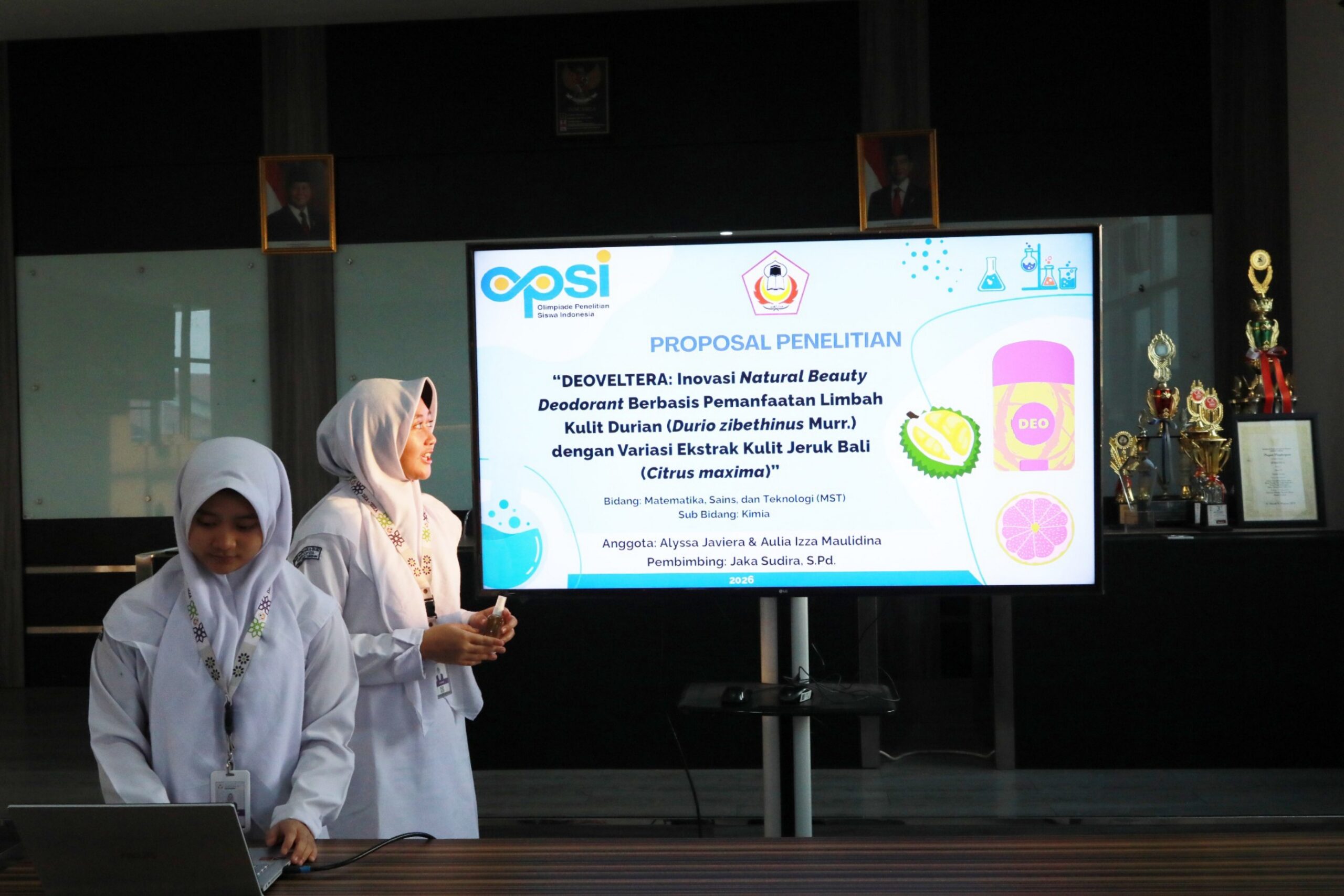 Two female students in white hijabs presenting a research poster on a screen about durian-based natural deodorant.