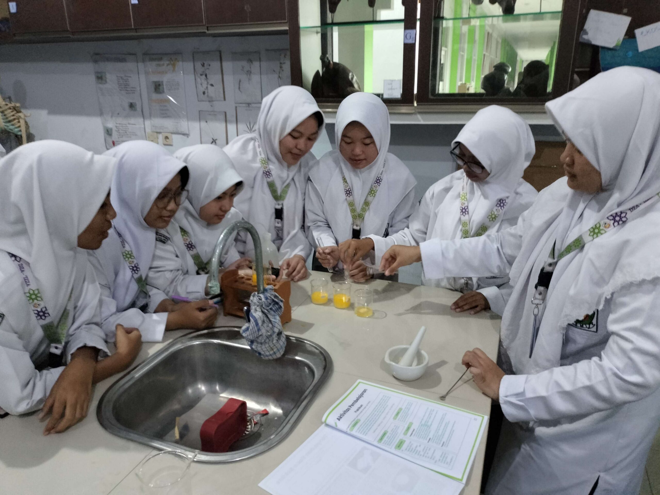 Seven female students in white lab coats and hijabs gathered around a lab bench doing a small chemistry experiment with beakers.