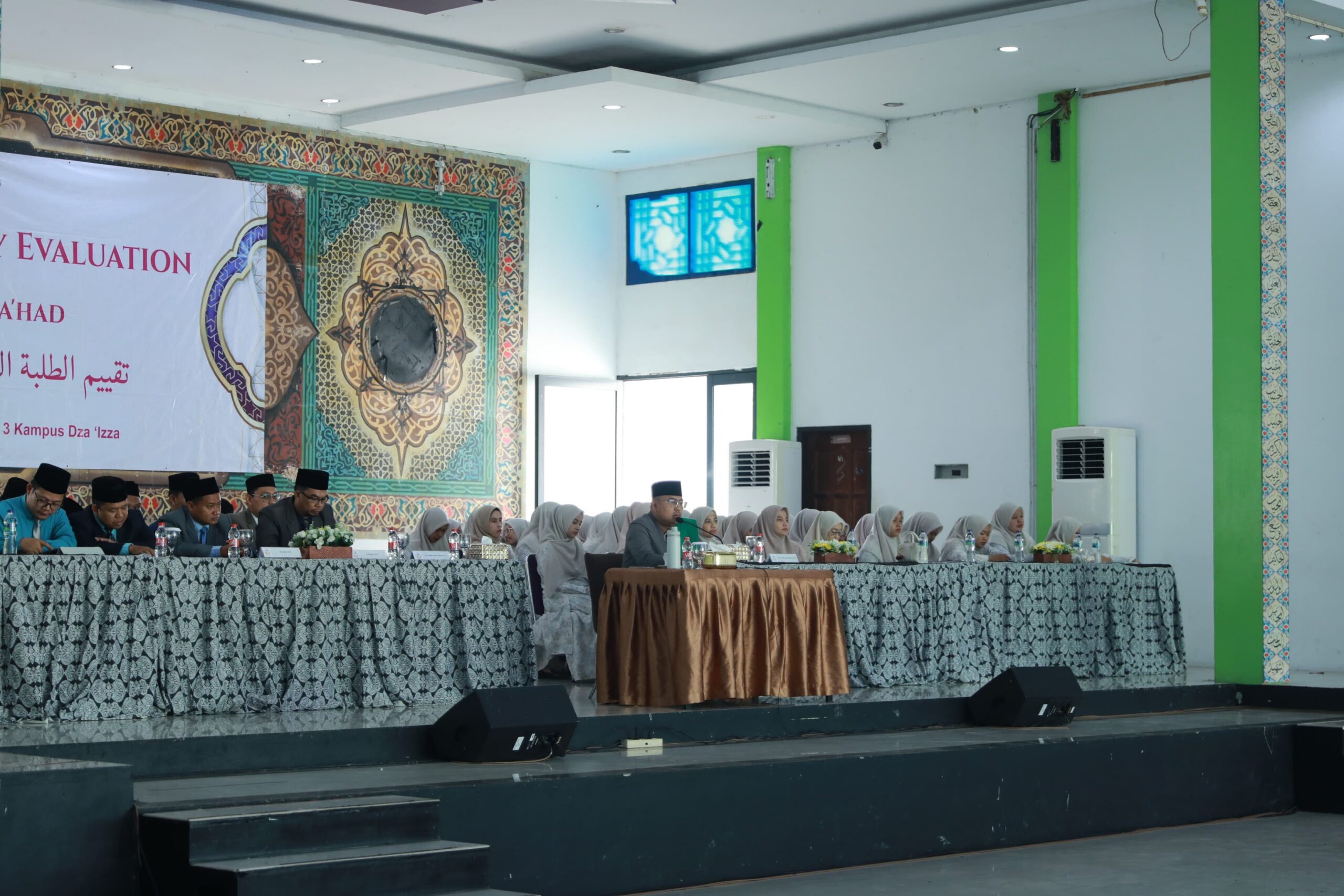 Long table on a stage with men in dark caps and women in light hijabs seated behind patterned cloths at a formal event.