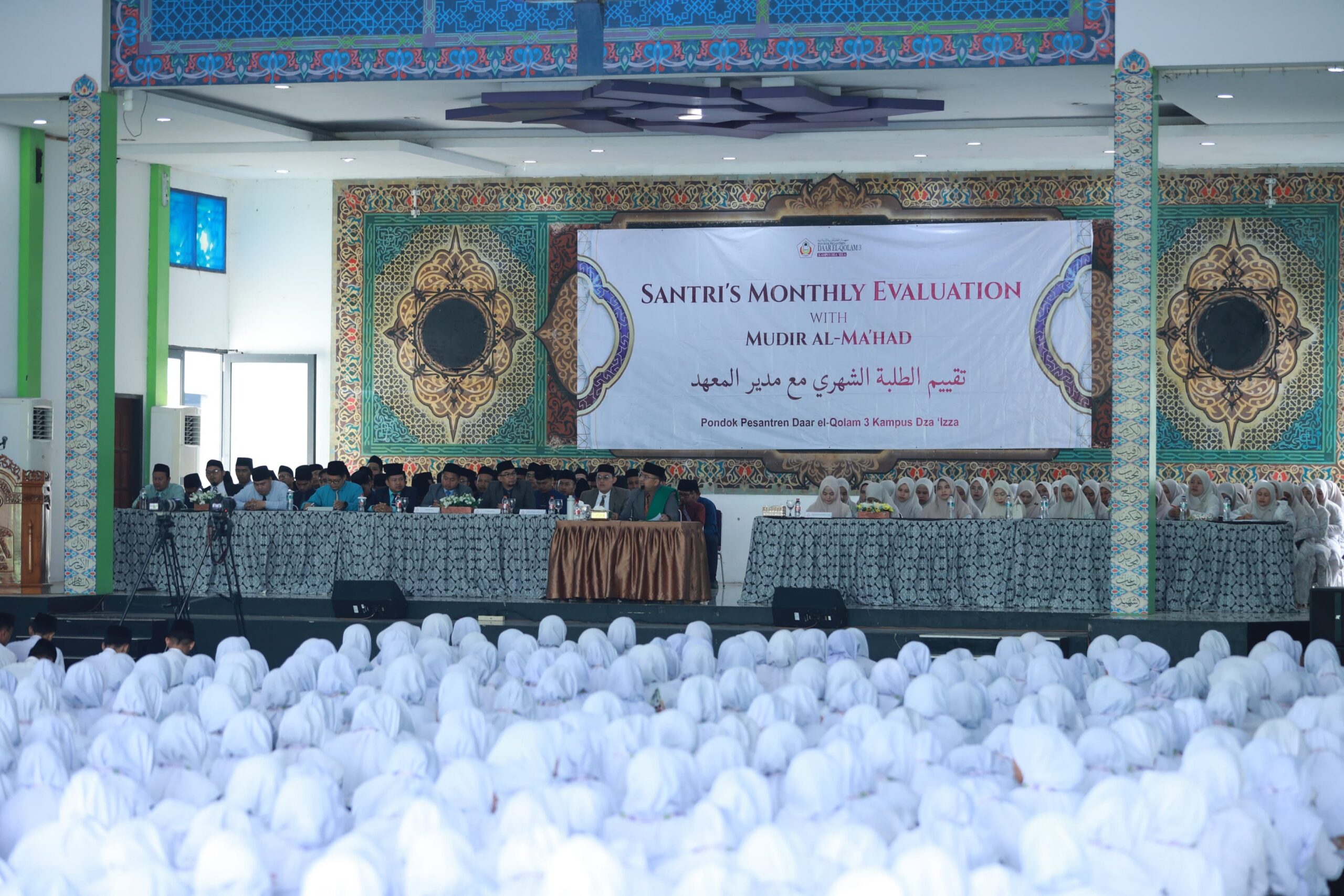 Large assembly of students in white headscarves facing a decorated stage where officials sit beneath a banner reading "Santri's Monthly Evaluation."