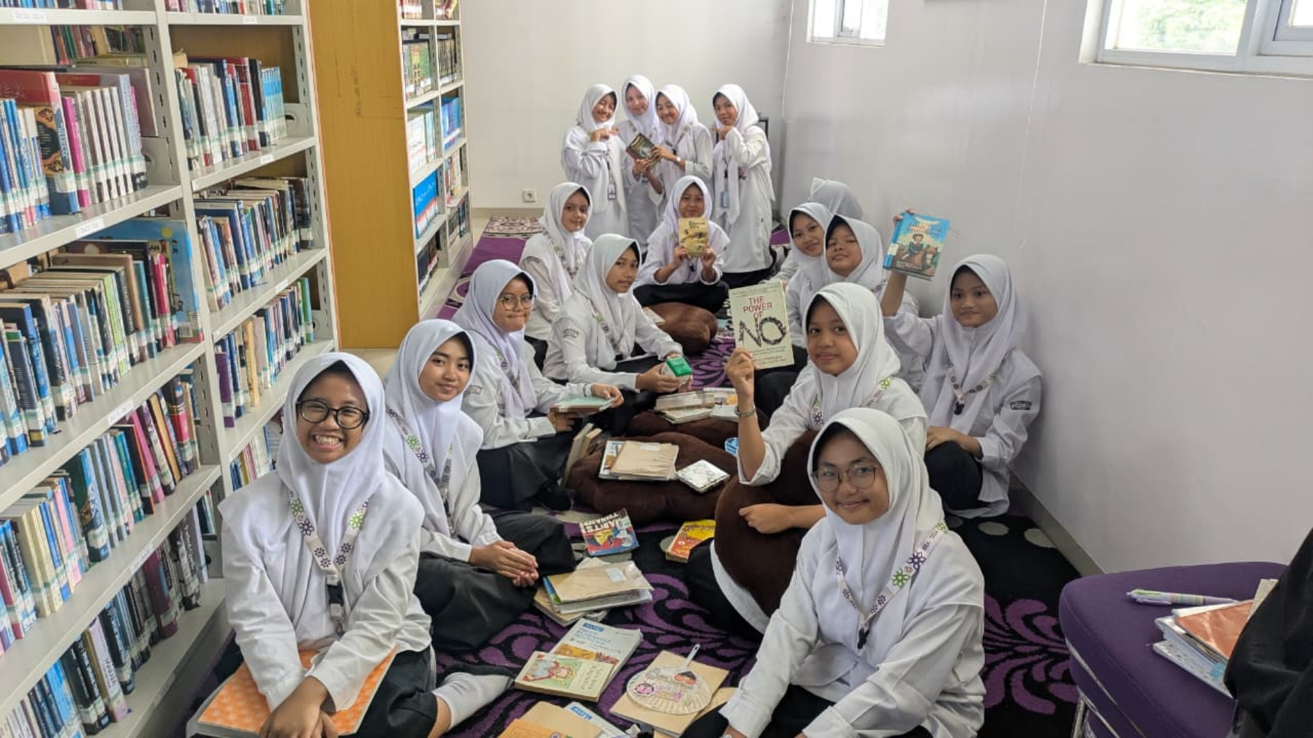 Group of schoolgirls in white hijabs sitting on a carpeted library floor, smiling and holding books with shelves visible along the left wall.
