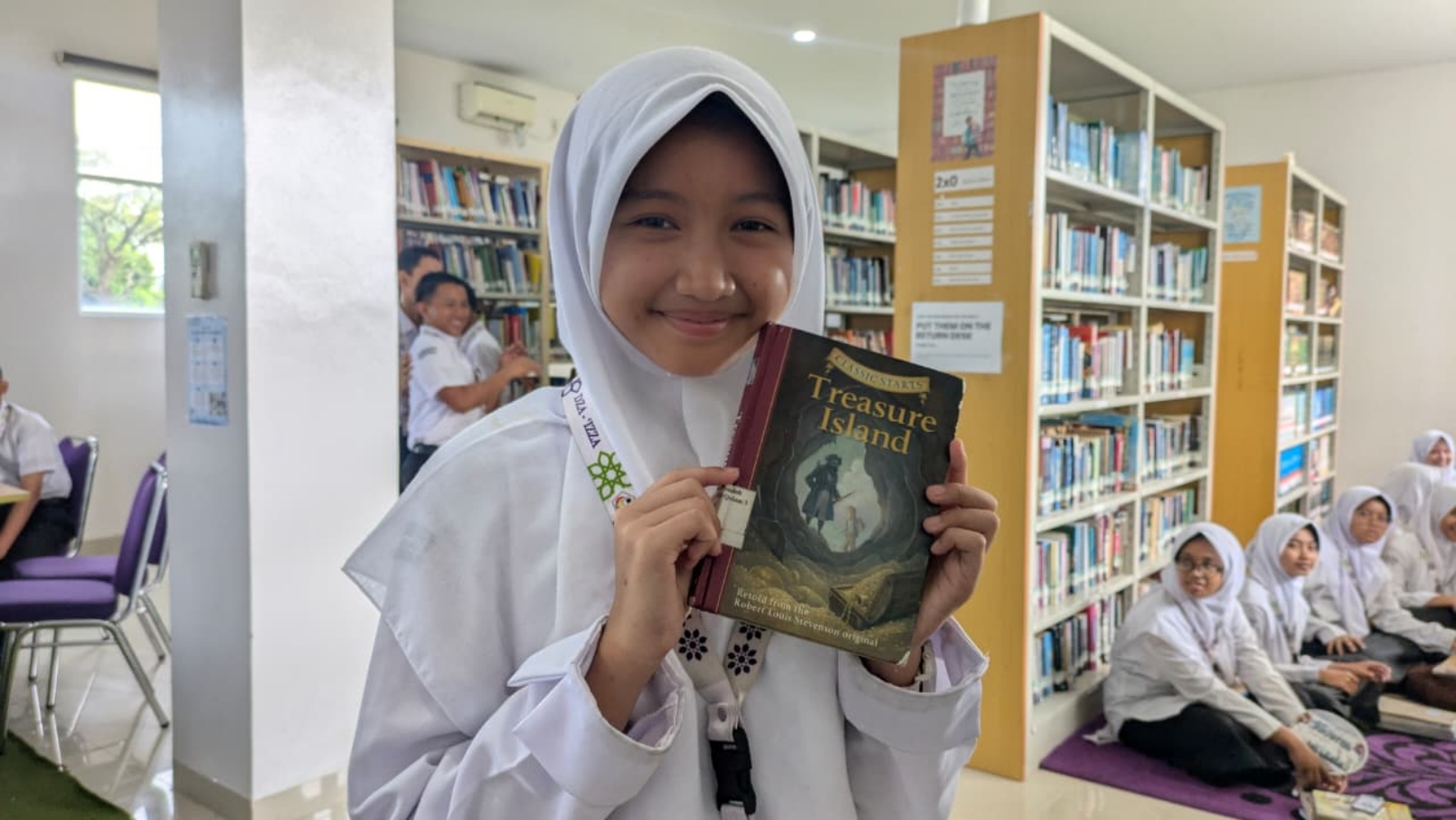 Smiling schoolgirl in a white hijab holding a worn copy of "Treasure Island" inside a library with bookshelves and seated classmates in the background