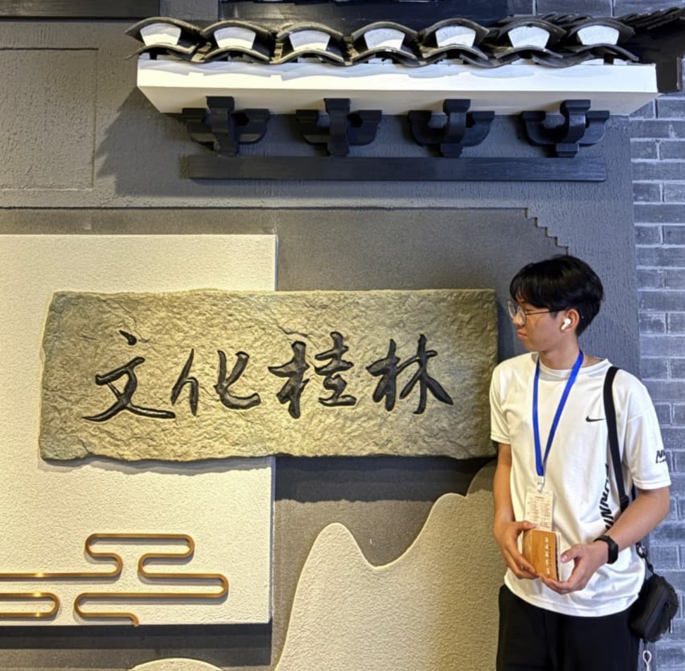 Teenage boy wearing glasses and earbuds stands beside a stone plaque with four Chinese characters on a textured wall.