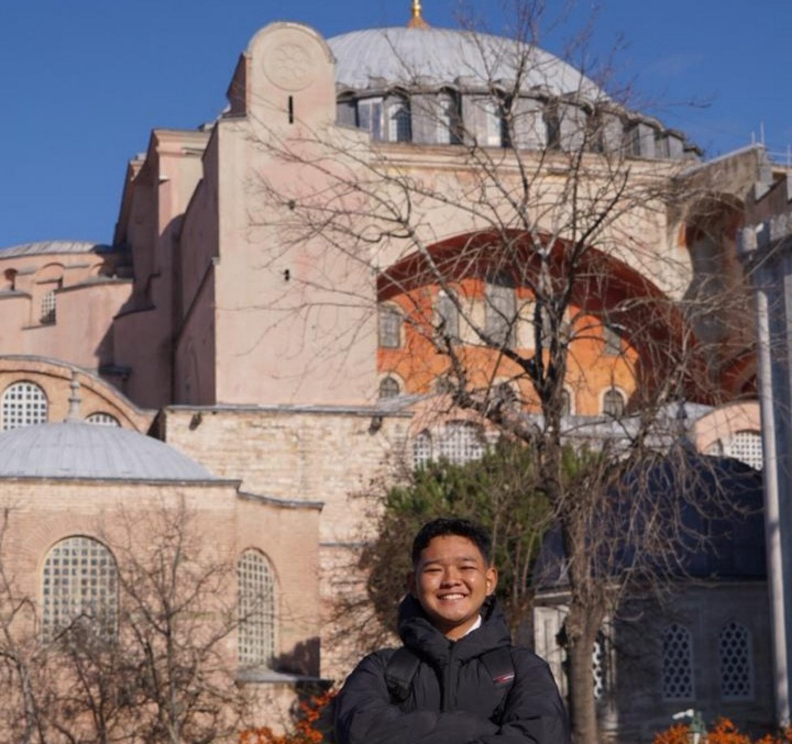 Smiling person in a black puffer jacket standing in front of a large historic domed building with bare trees in foreground