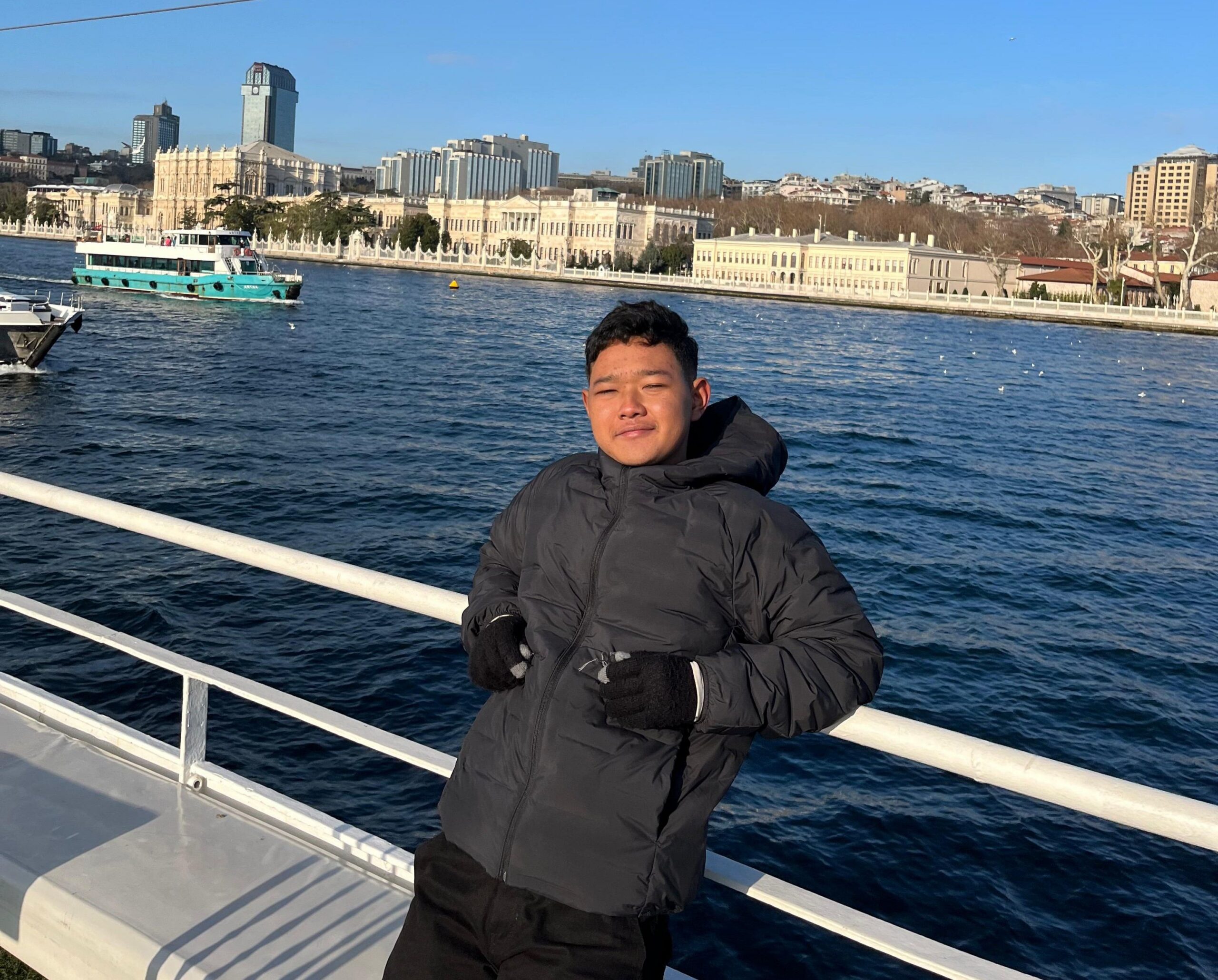 Young man in a black puffer jacket leaning on a white railing with a ferry and city skyline across blue water