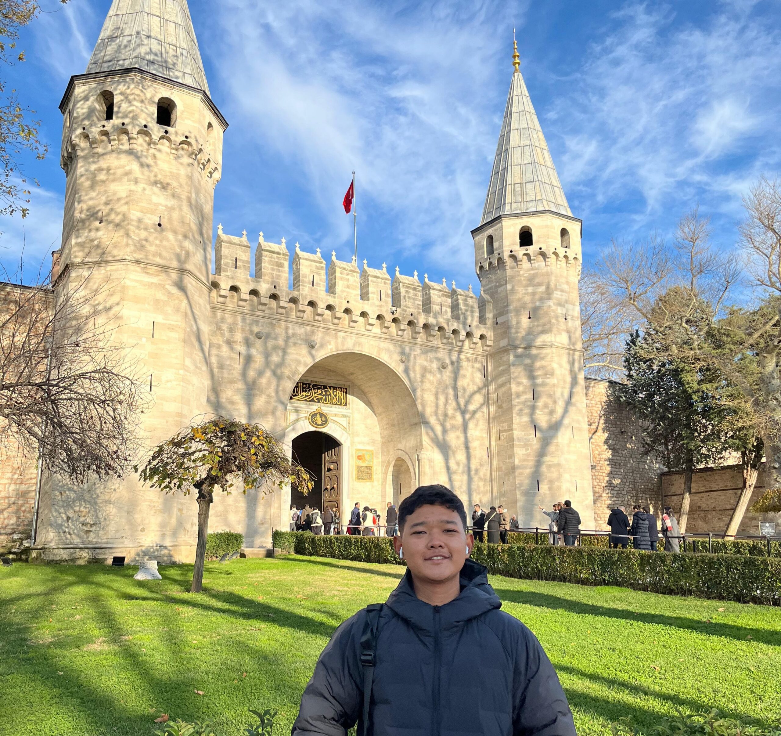 Young person in a dark puffer jacket smiling in front of a grand stone gate with twin conical towers and a Turkish flag.