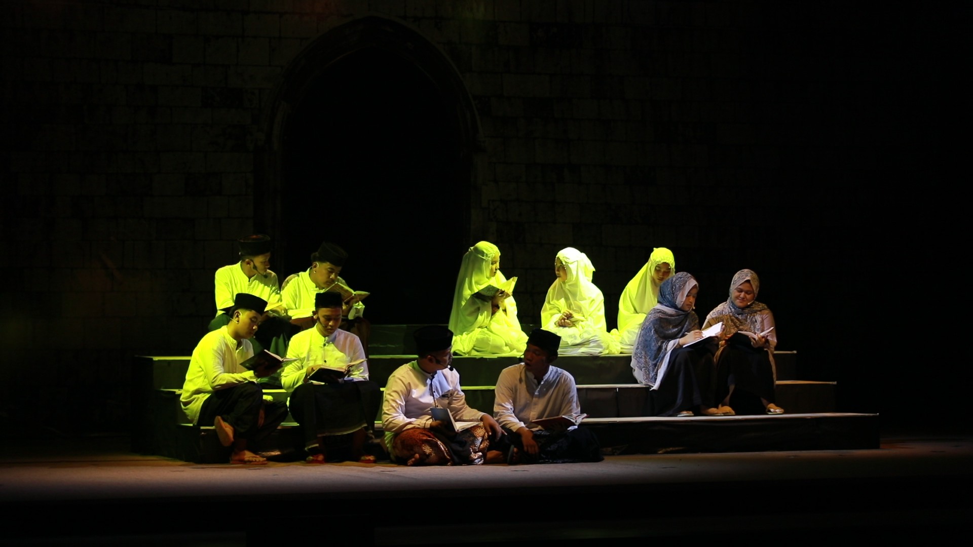 Group of men and women in traditional Muslim dress seated on tiered stage steps, reading books under warm yellow spotlight against dark brick backdrop.