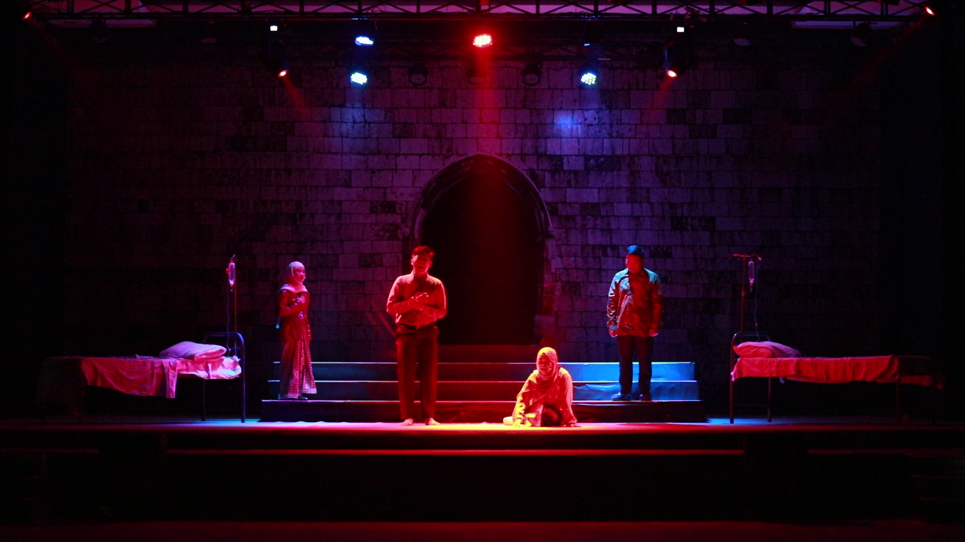 Four actors on a dim stage lit in red and blue, two hospital beds at sides, one actor kneeling centre with a stone arch backdrop.