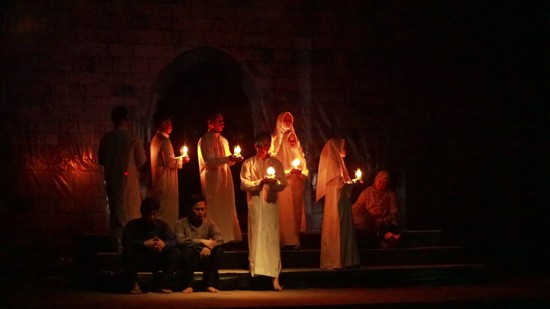 Group of robed men and women holding lit candles in a dark, arched stage setting, faces softly illuminated by the flames