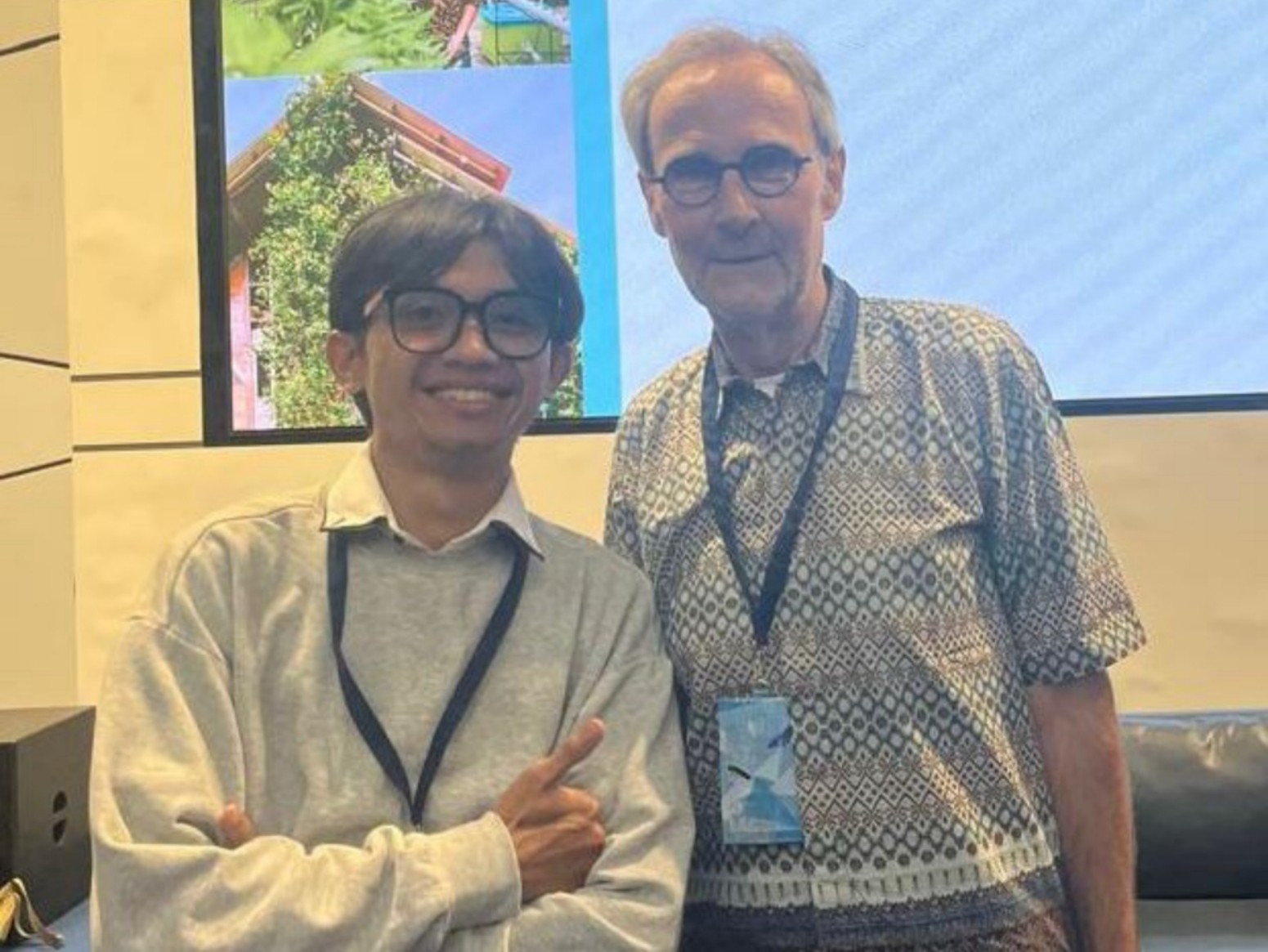 Two smiling men wearing glasses and lanyards standing side by side in front of a large screen, one in a grey sweater and the other in a patterned short-sleeve shirt.