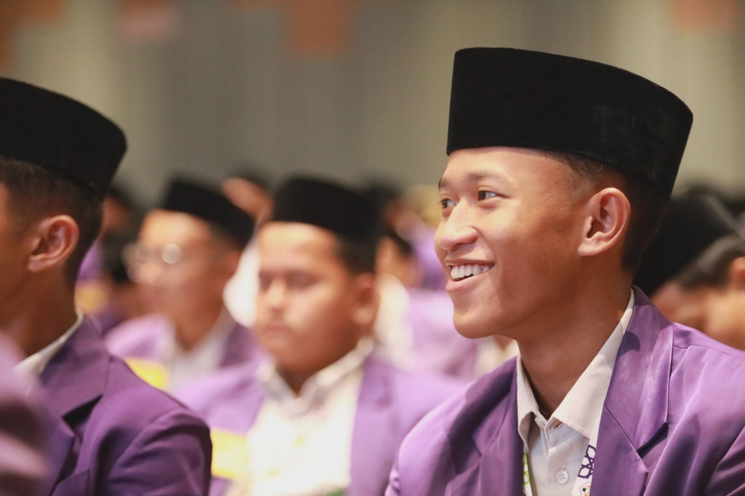 Smiling young man in a black songkok and purple uniform jacket seated among peers at a formal event