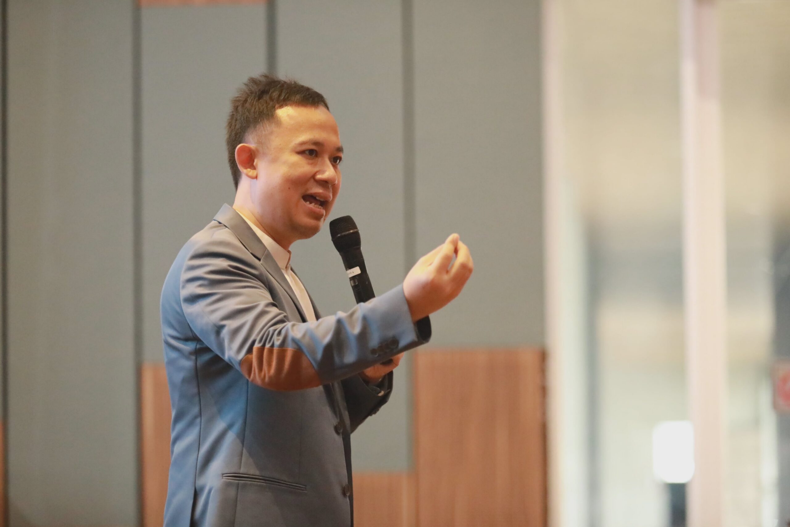 Man in a grey blazer speaking into a handheld microphone, gesturing with his right hand during a presentation.