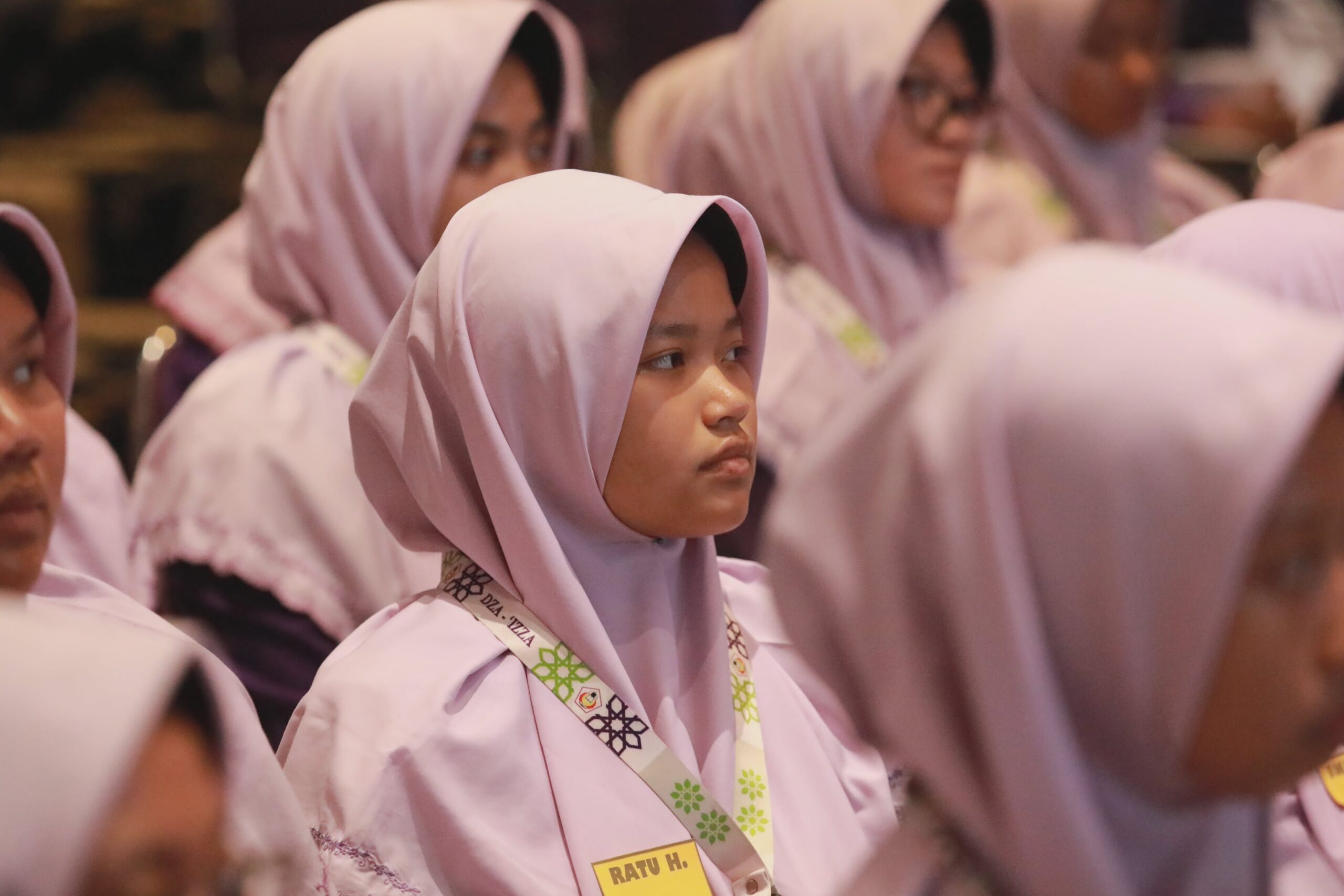 Teen girl in a pale lilac hijab and lanyard looking to the side among other girls in similar headscarves.