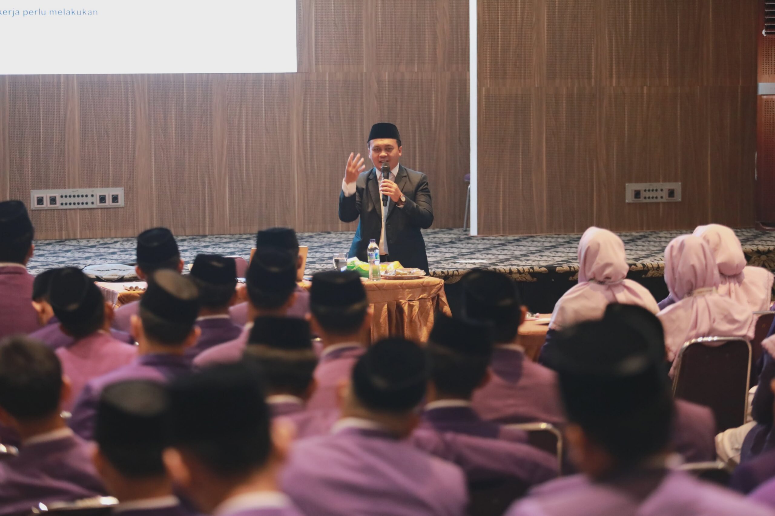 Man in suit and songkok speaking into a microphone at front of a conference room with audience in purple uniforms.