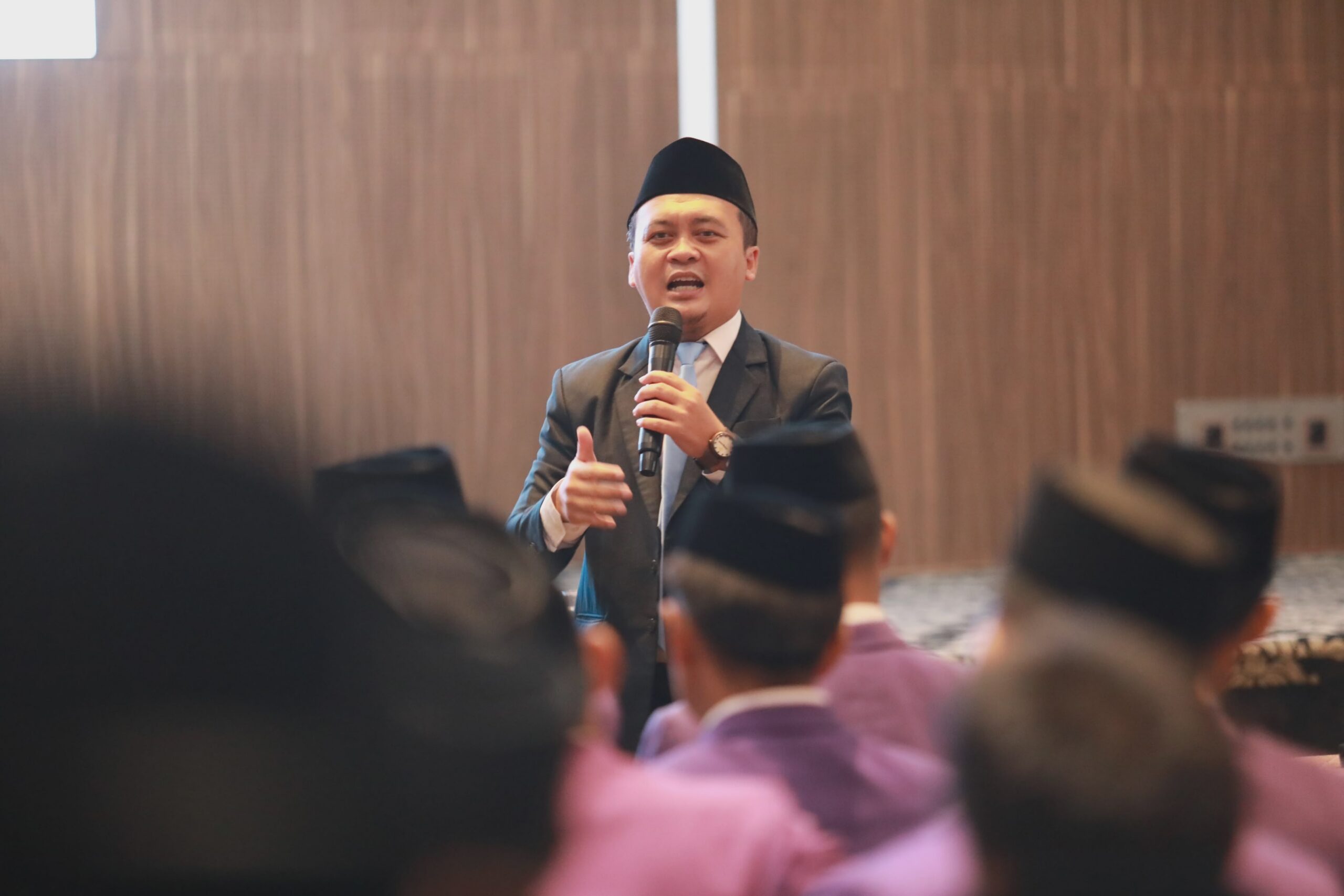 Man in a suit and traditional black cap speaking into a microphone to an audience, gesturing with his hand.