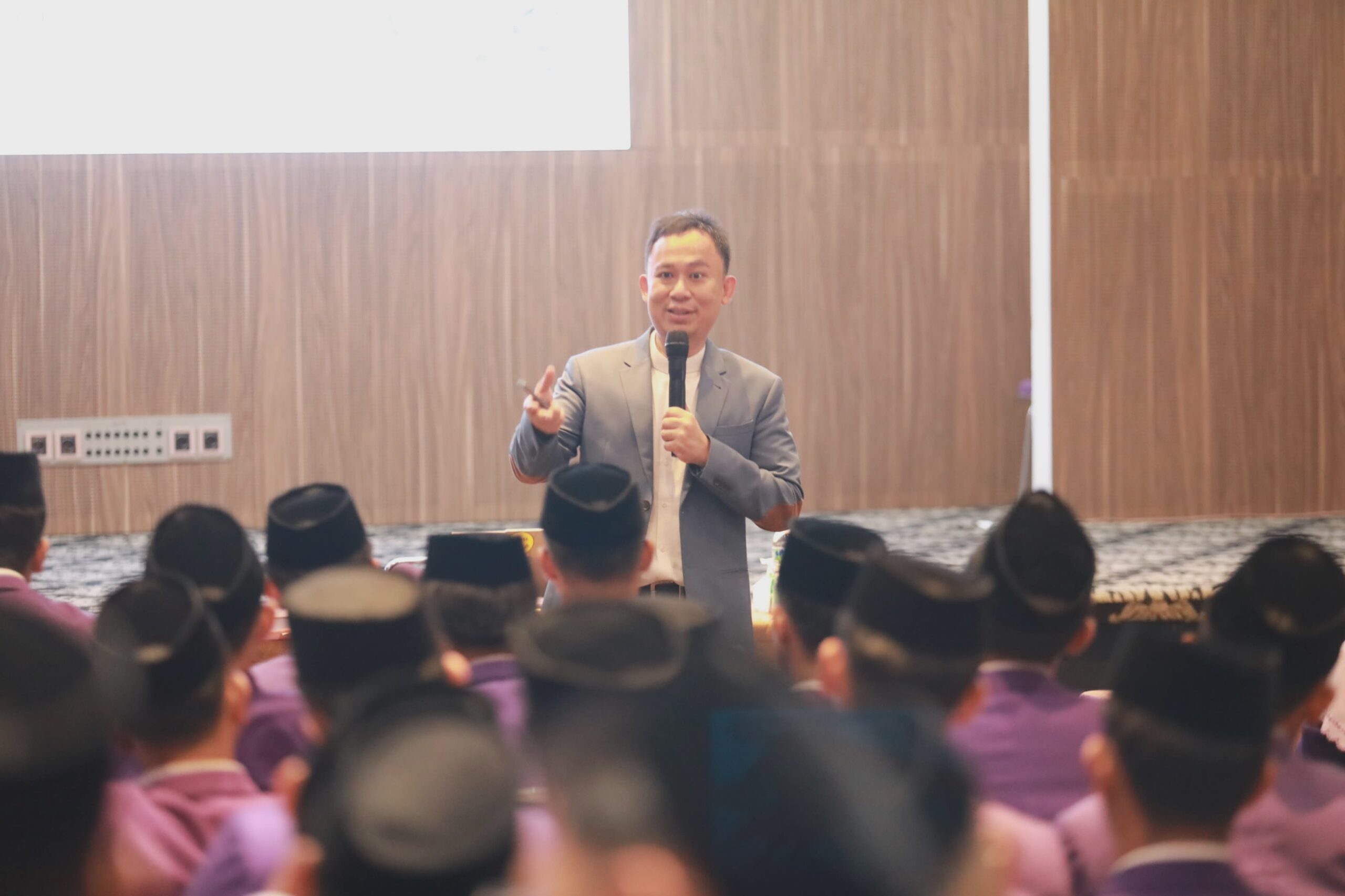 Man in a suit speaking into a microphone to an audience of seated people wearing black caps in a conference room