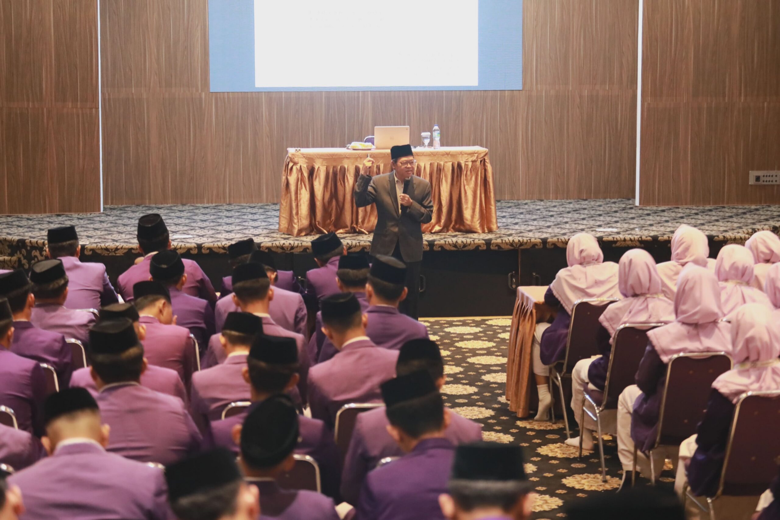 Man in suit and cap speaking to seated audience in purple uniforms and headscarves at a conference room lectern.