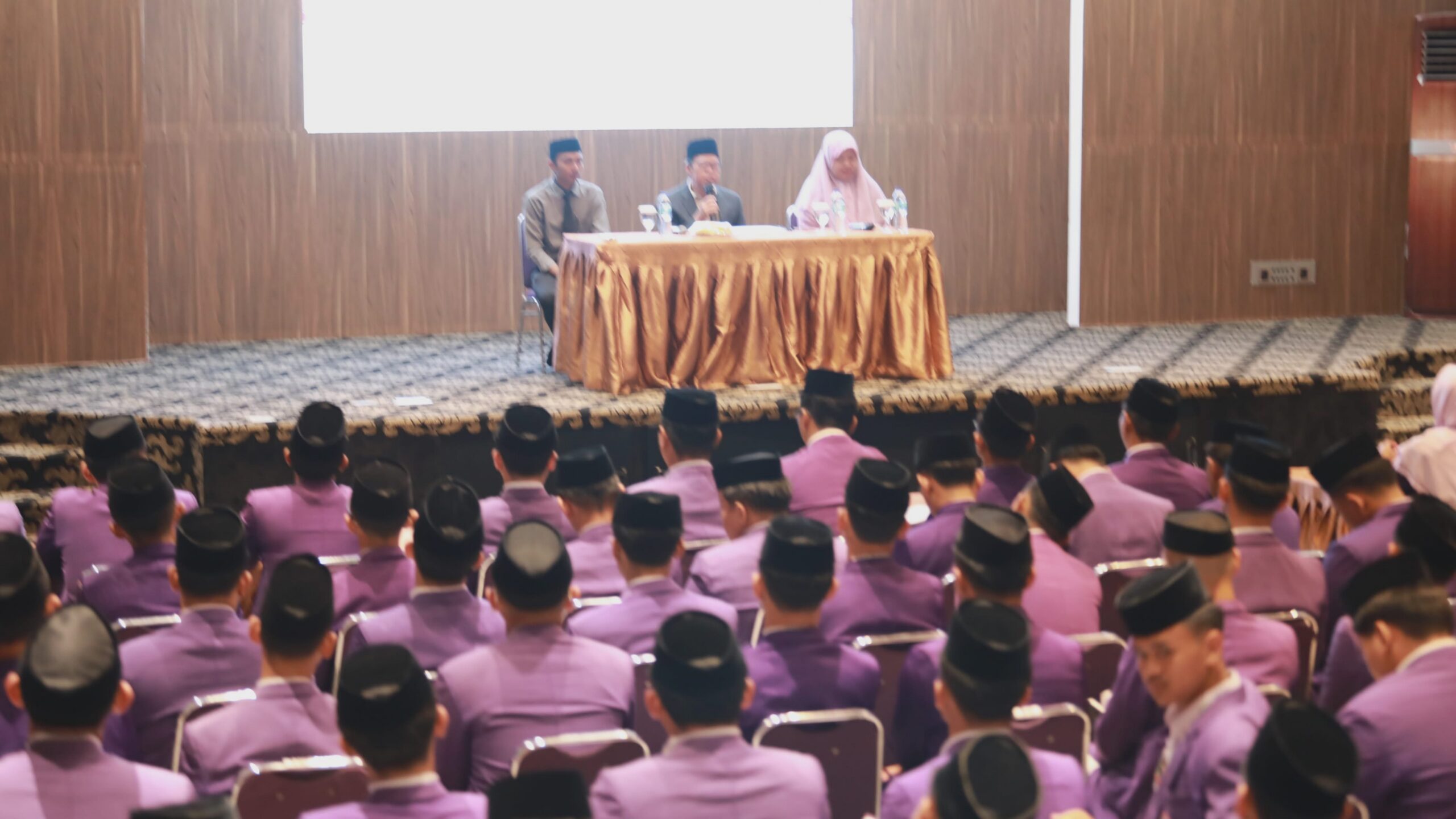 Audience in purple uniforms and black caps seated facing a panel of three speakers at a draped table on stage.