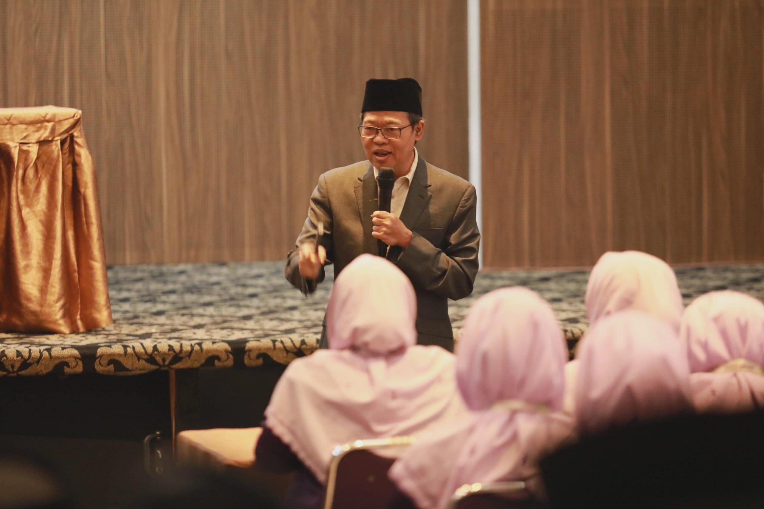 Man in suit and black cap speaking into a microphone to an audience of women wearing pink headscarves in a conference room