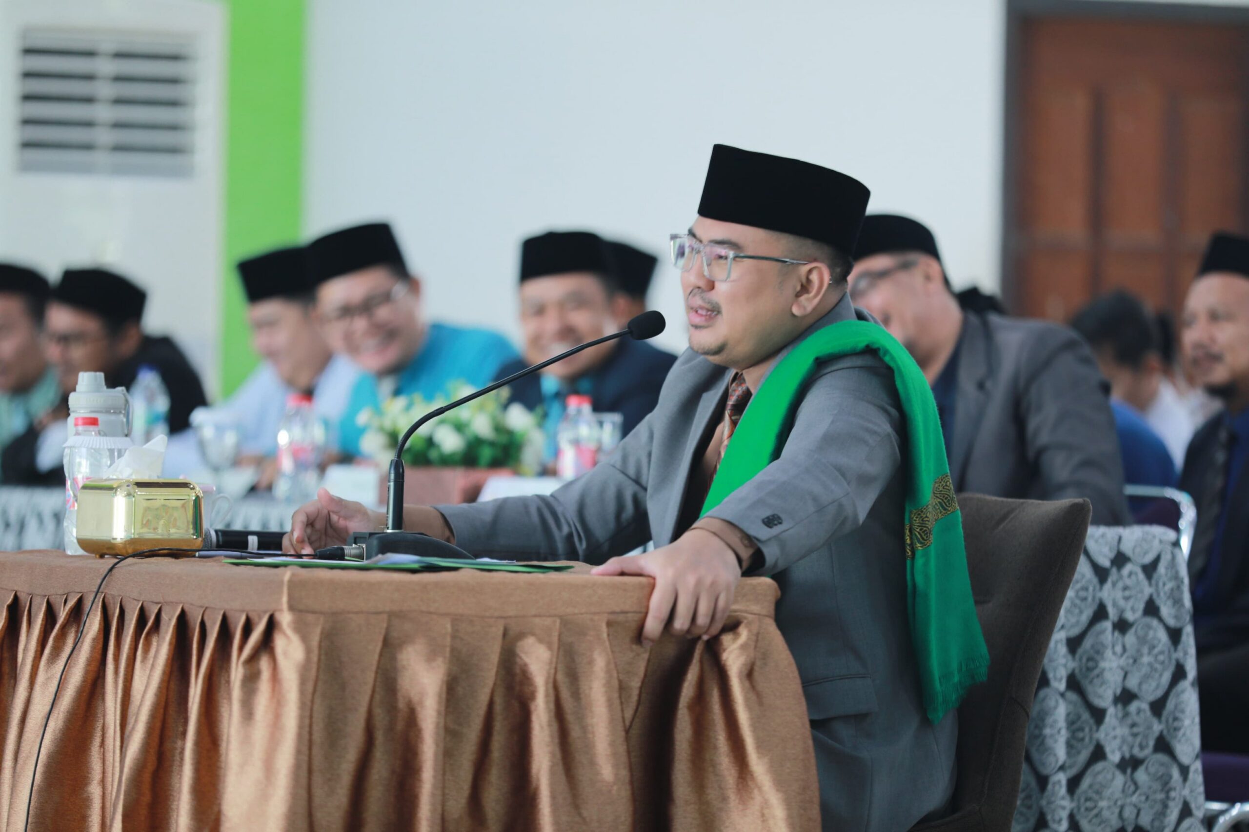 Man in traditional cap and glasses speaking into a microphone at a conference table, wearing a green sash and grey suit.