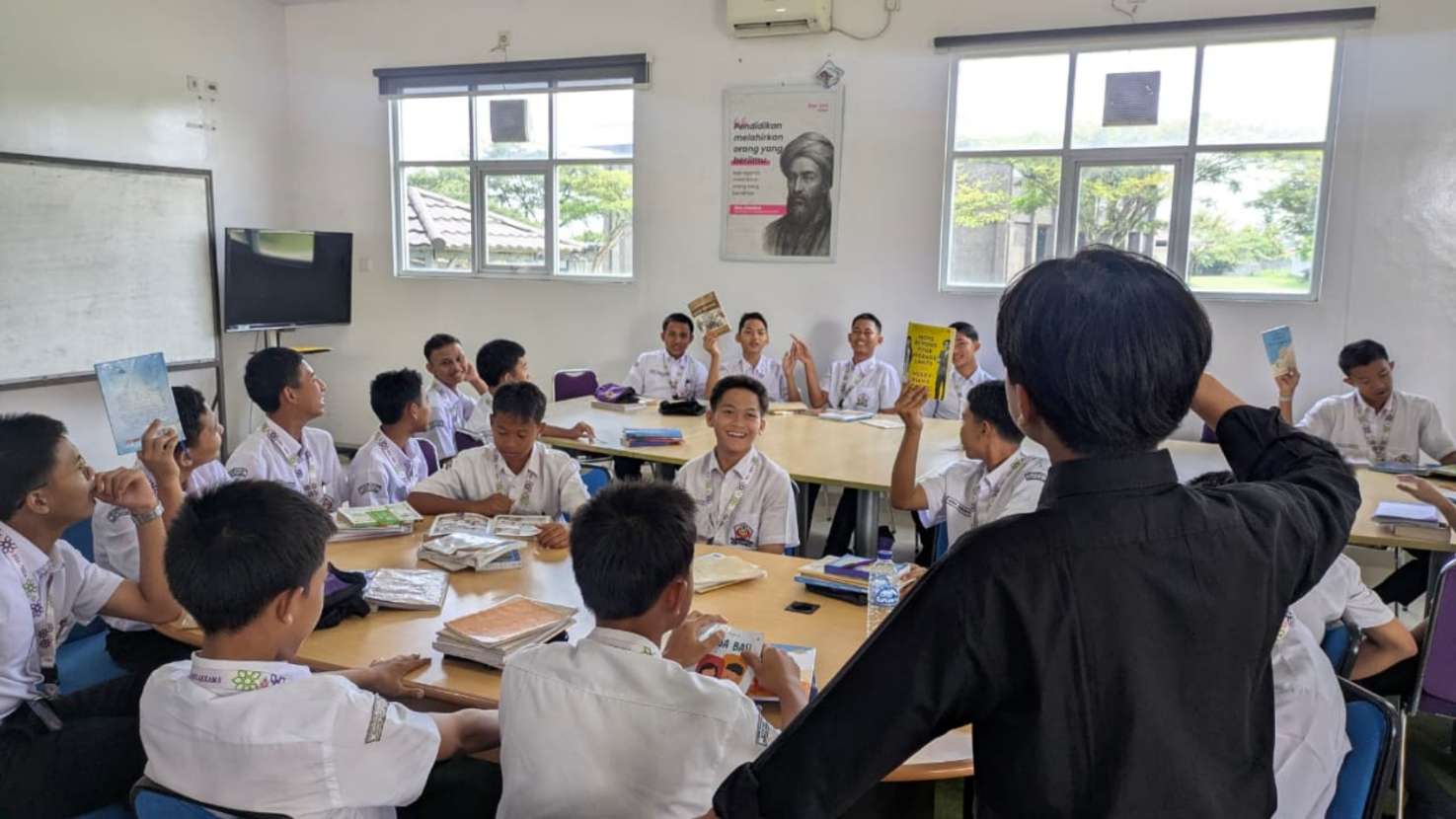Teenage boys in school uniforms seated around tables in a bright classroom, some holding up books while a student stands speaking.