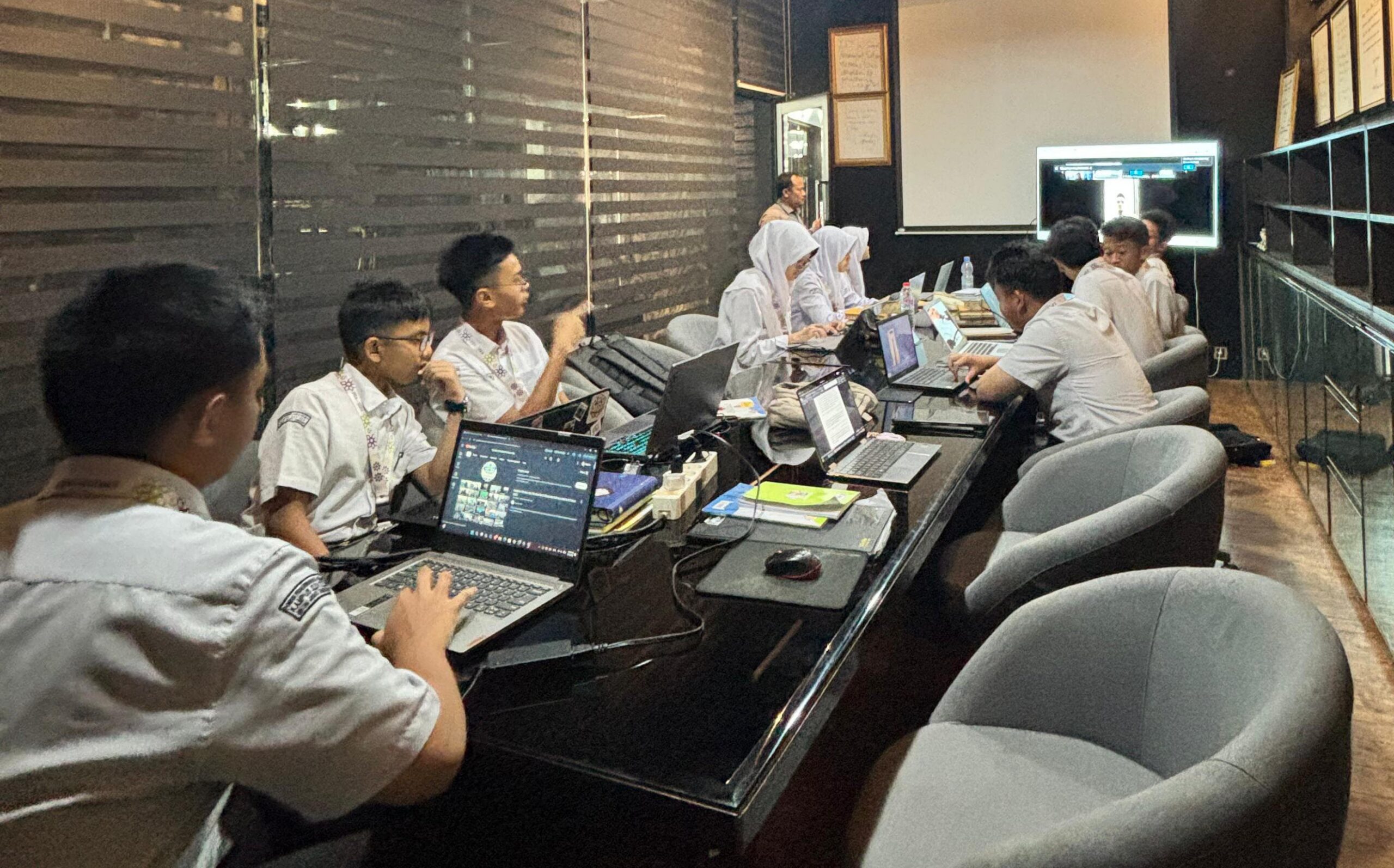 Students in school uniforms sit around a long table using laptops while a teacher stands near a projector screen.