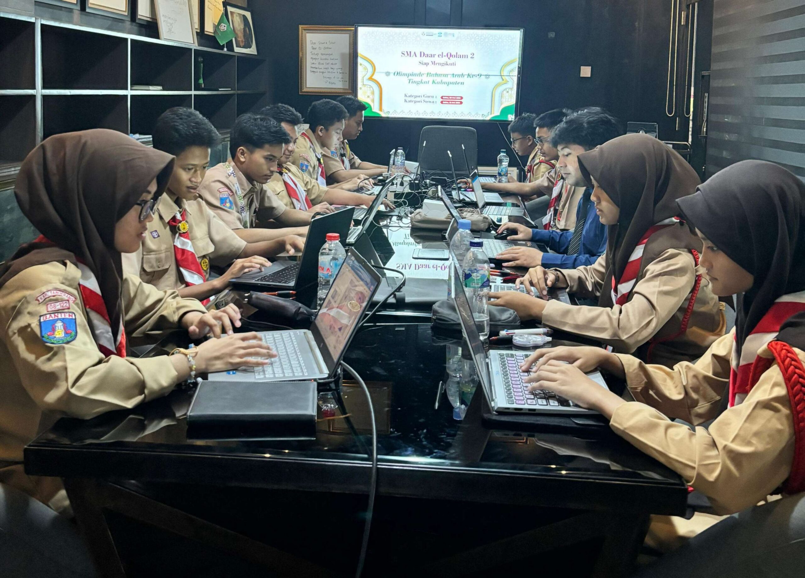 Teen scouts in uniform and headscarves seated along both sides of a long table, working on laptops in a meeting room