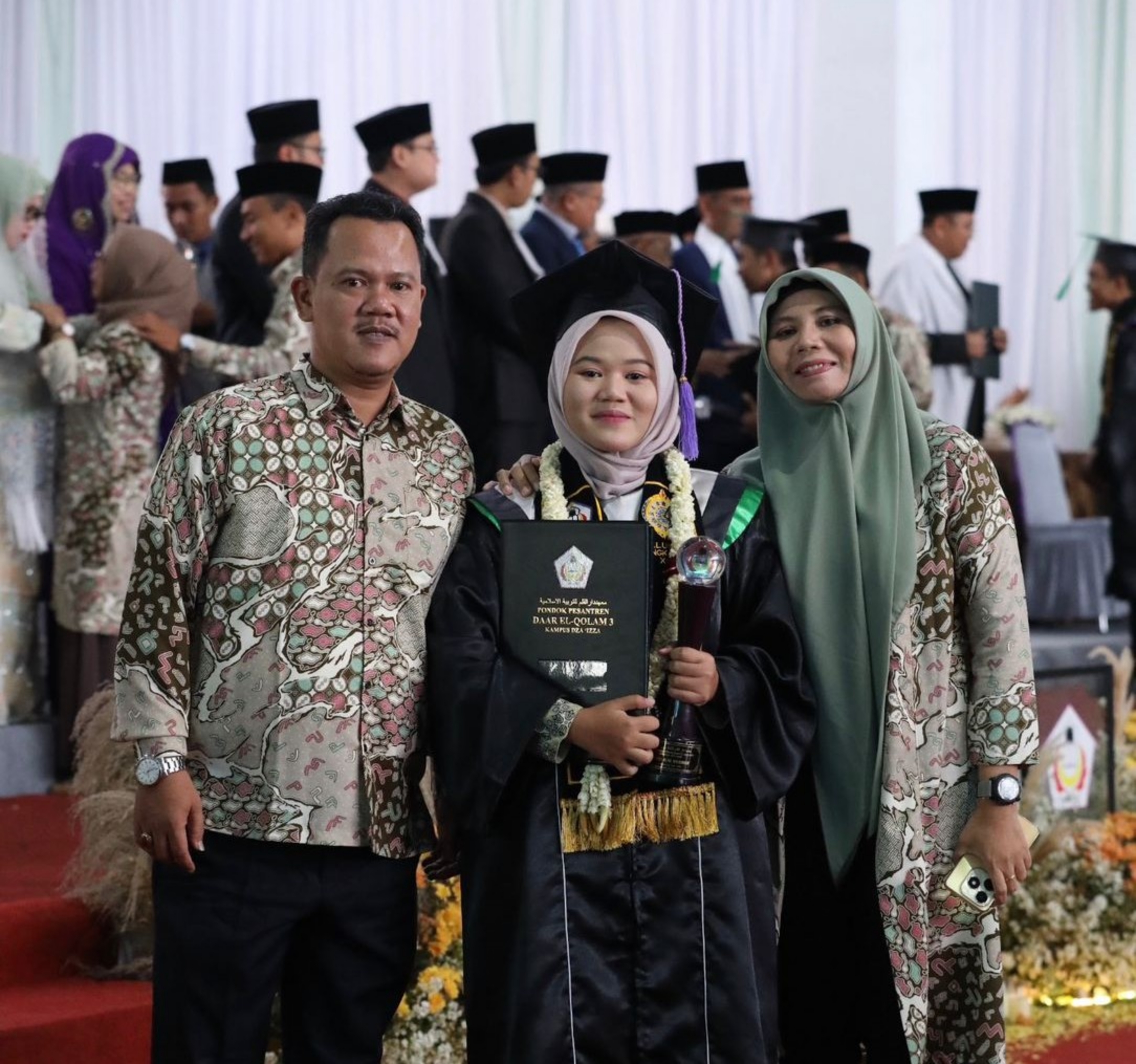 Young graduate in cap and gown holding diploma and trophy, flanked by proud parents wearing patterned batik and hijab.
