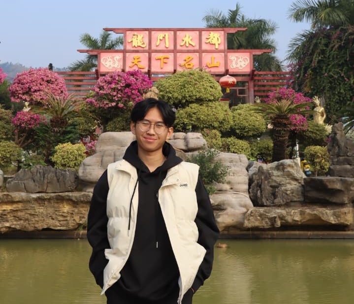 Young man wearing glasses and a light vest standing with hands in pockets in front of a pond, rock garden and colourful flowering shrubs with a red Chinese signboard behind.