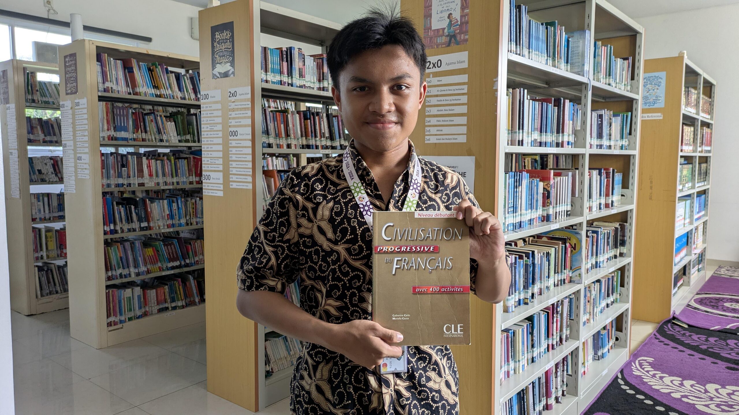 Teenage boy in a patterned shirt standing in a library aisle holding a French textbook titled "Civilisation progressive du français."