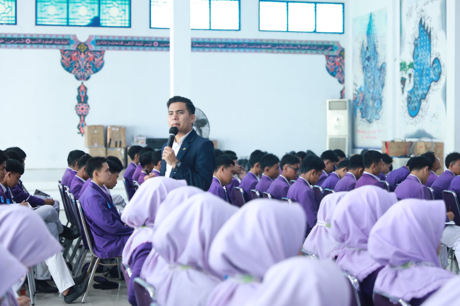 Male speaker with microphone addressing rows of students in purple uniforms seated in a hall, some girls wearing light purple hijabs in foreground.