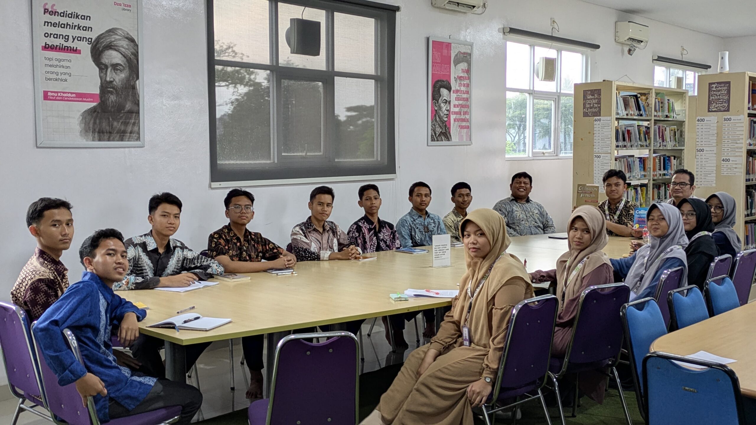 Students and teacher seated around a long table in a bright library, many wearing batik shirts and hijabs, bookshelves behind them