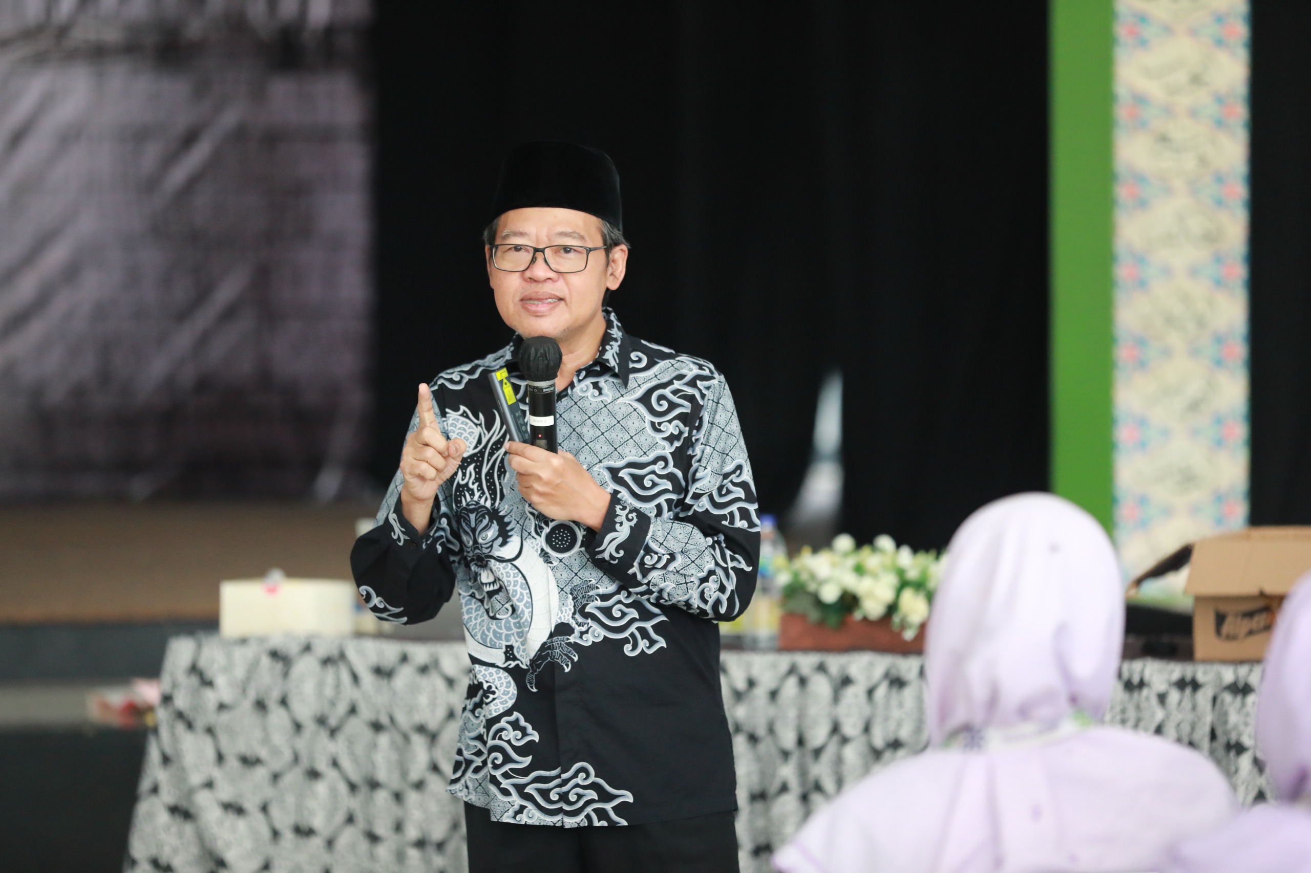 Middle-aged man in glasses and a patterned black‑and‑white batik shirt and songkok speaking into a microphone, gesturing with his finger.