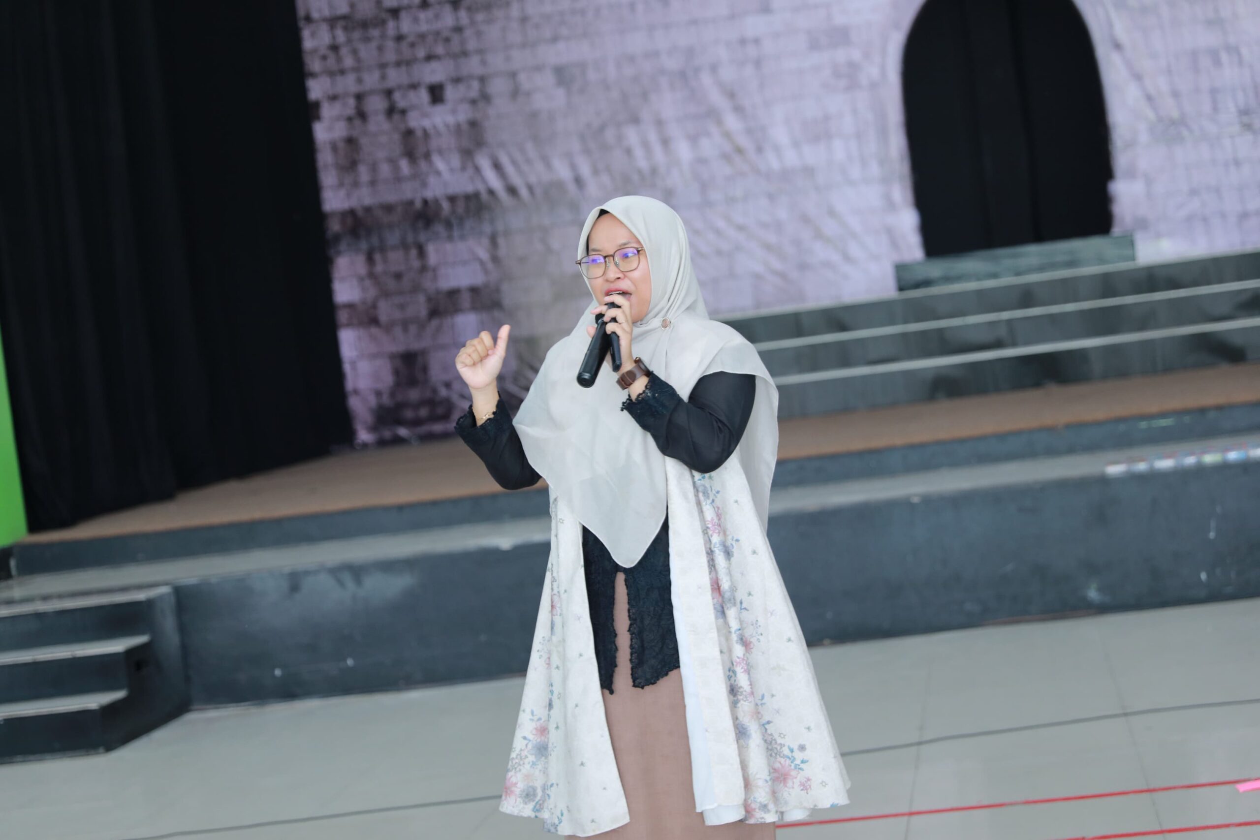 Woman wearing a pale hijab and glasses speaking into a microphone on a stage with steps behind her