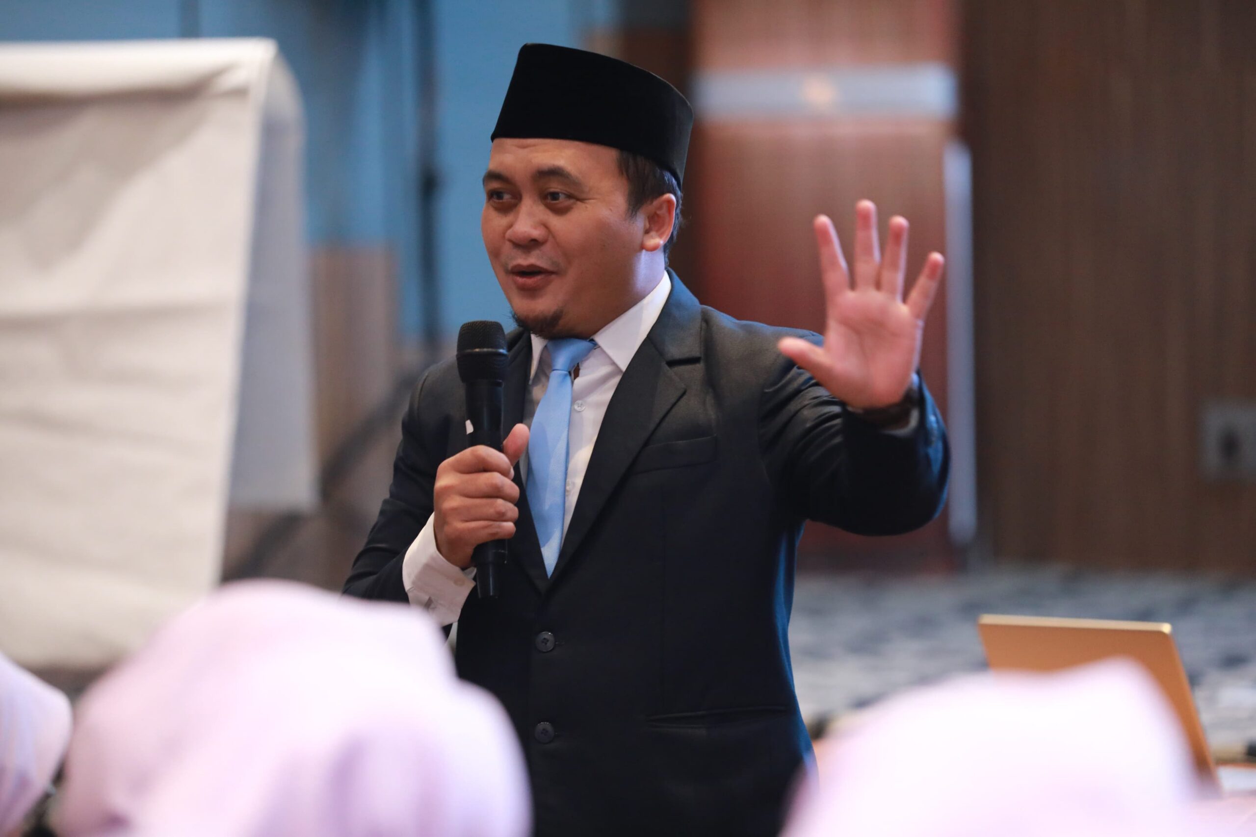 Man in suit and traditional black cap speaking into a microphone and raising his hand while addressing an audience.