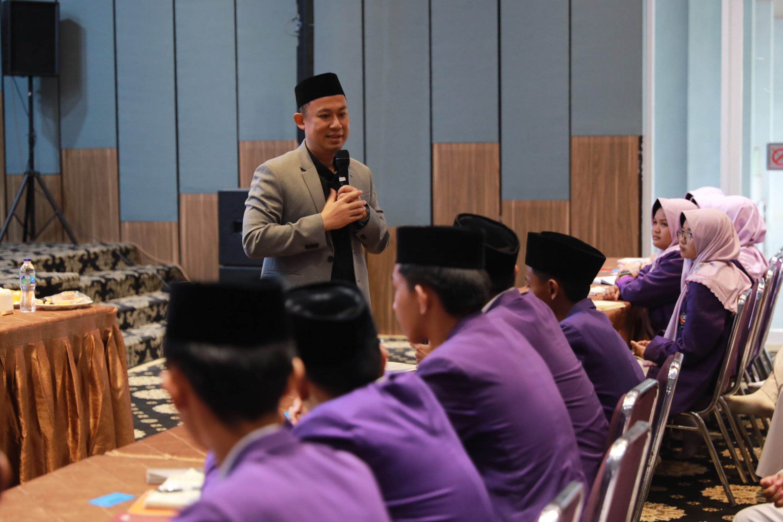 Man in grey blazer and black cap speaking into a microphone to seated students in purple uniforms and headscarves.