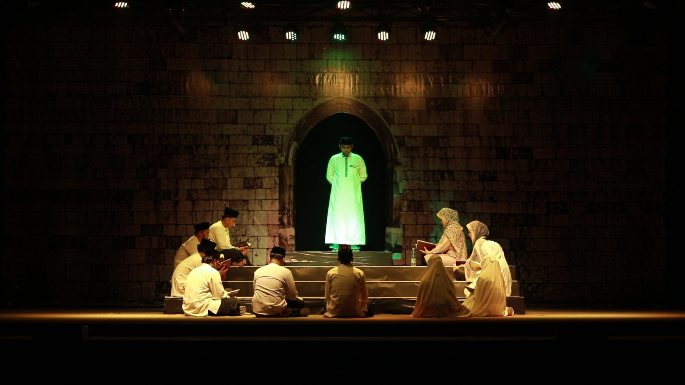Man in a green-lit robe stands in an arched doorway while a small group kneels and reads aloud on a dim stage.