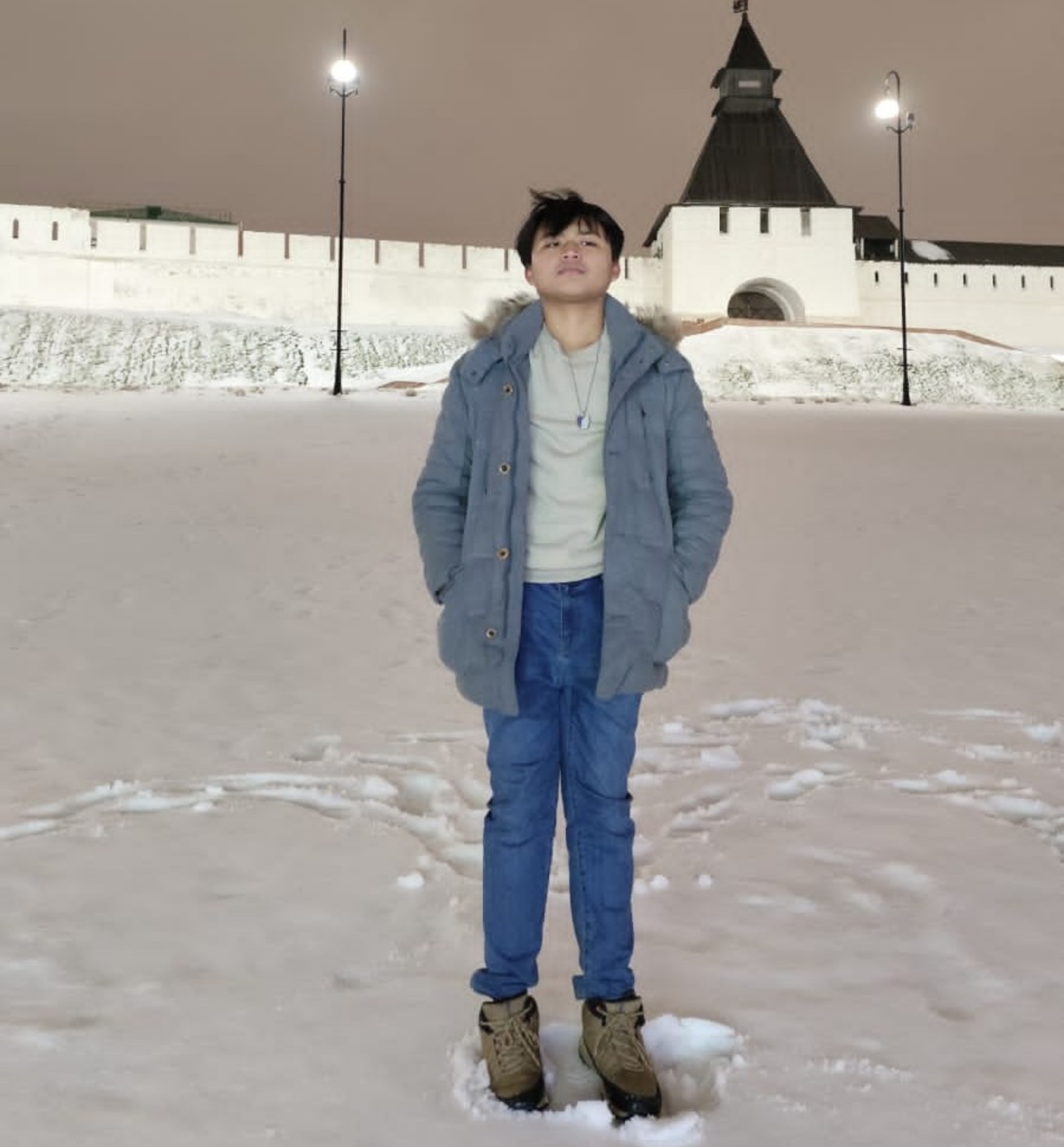 Teenage boy in a blue parka and jeans standing in snow with boots on, a lit fortress gate and lampposts in the background.