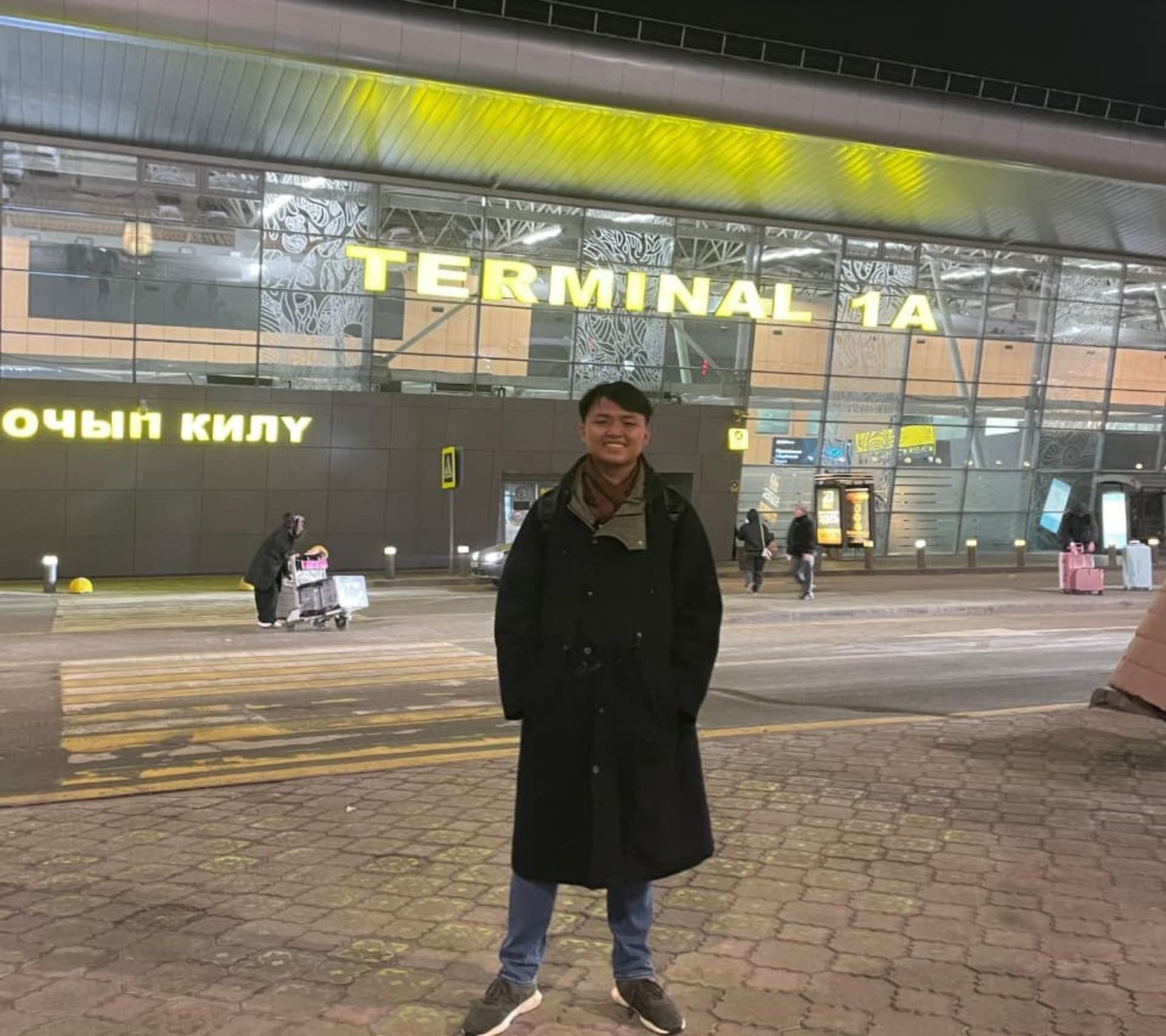 Person in a long dark coat standing outside a brightly lit airport entrance with large yellow "TERMINAL 1A" signage.