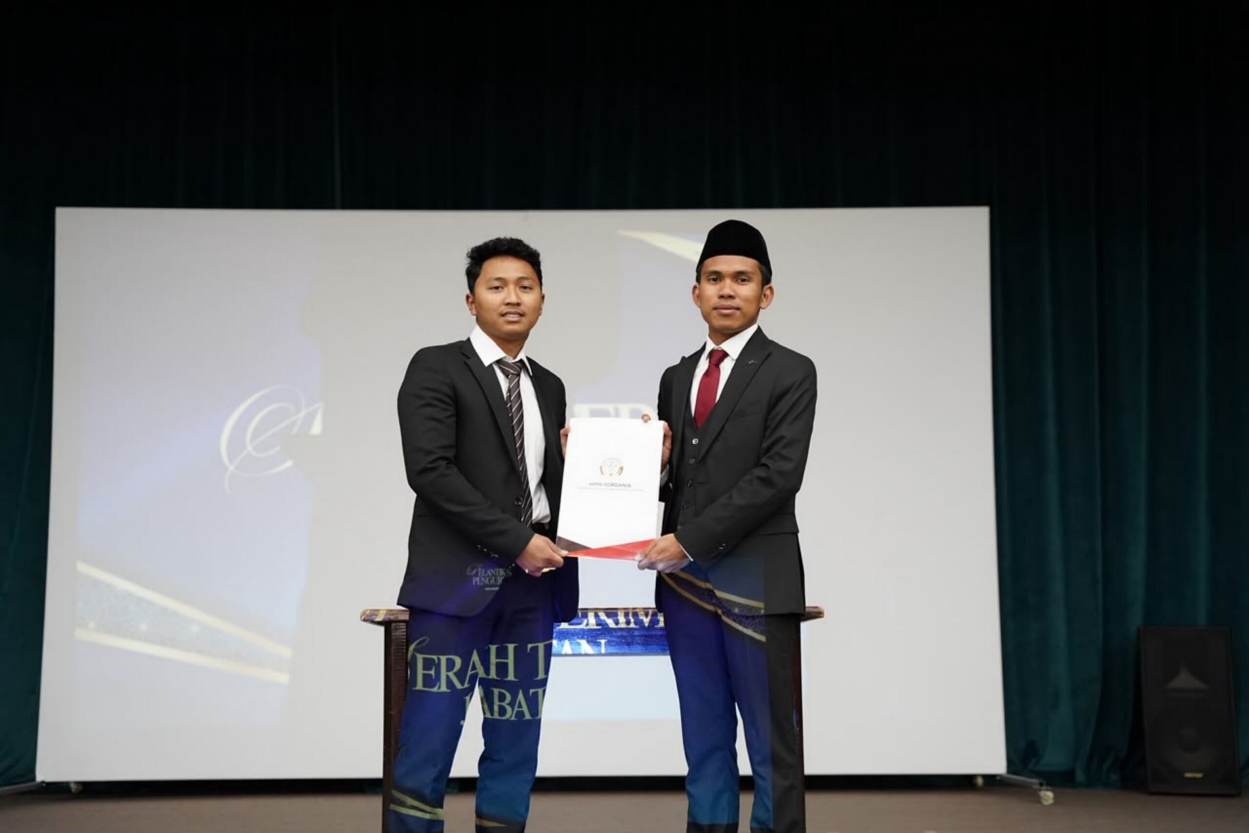 Two young men in suits standing on stage holding a certificate together, one wearing a black songkok hat.