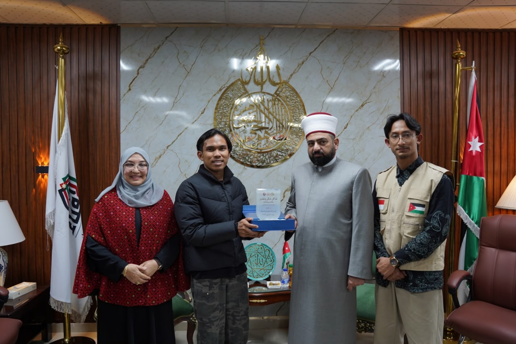 Four people standing in an office receiving a glass award, with a man in a grey religious robe and cap, flags and ornate emblem behind them.