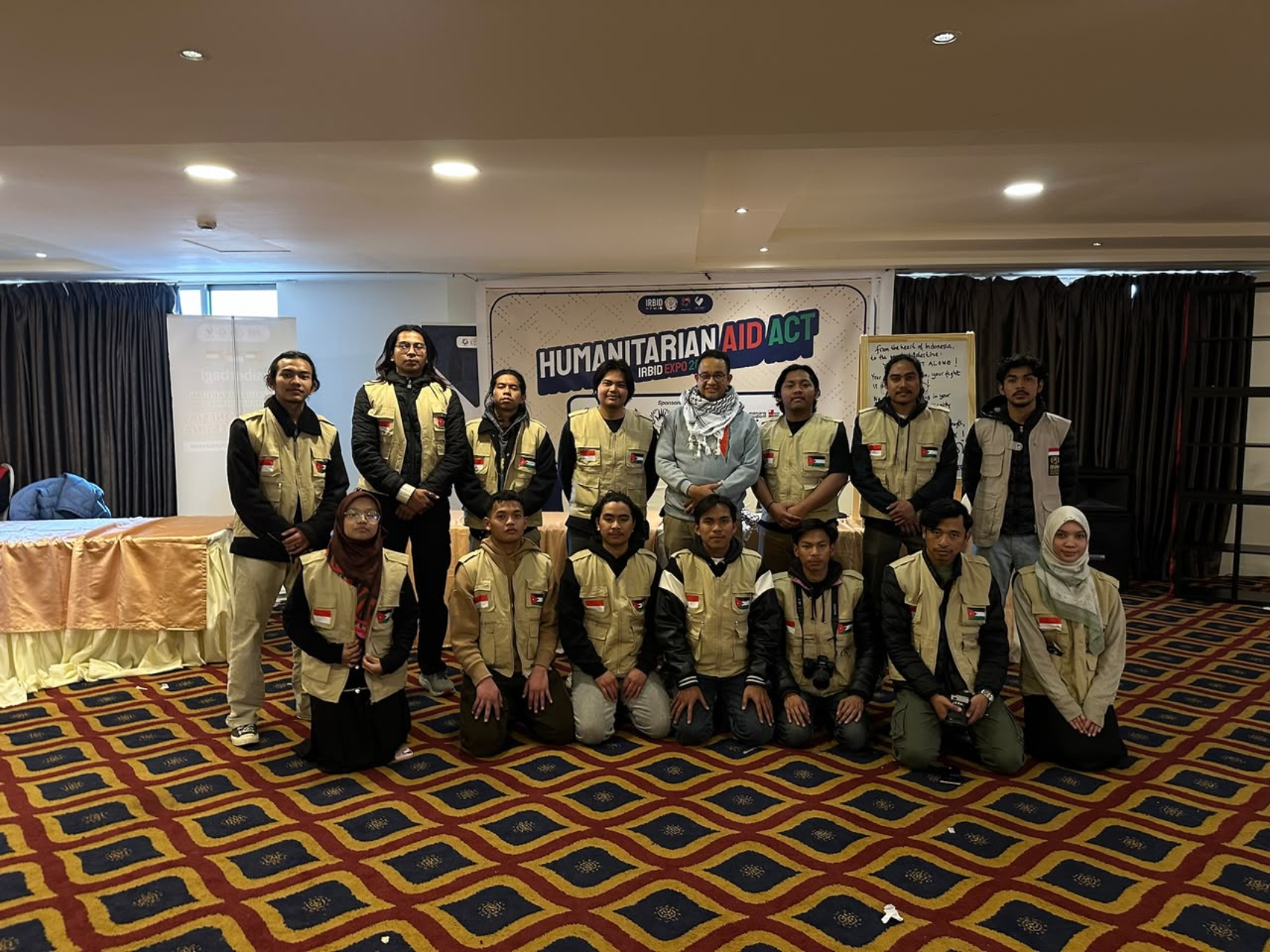 Group of volunteer aid workers wearing beige vests posing in a conference room in front of a "Humanitarian Aid Act" banner