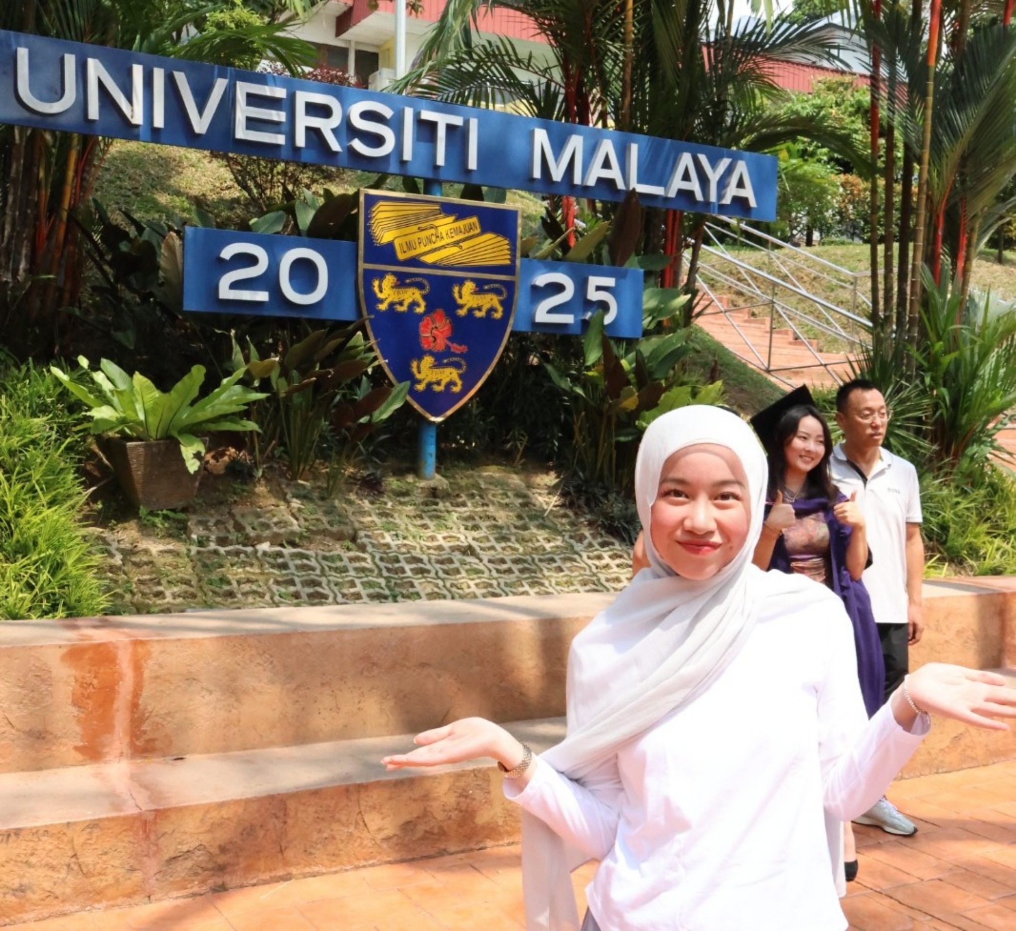 Smiling woman in a white hijab posing in front of a blue "Universiti Malaya" sign and crest with palm trees behind