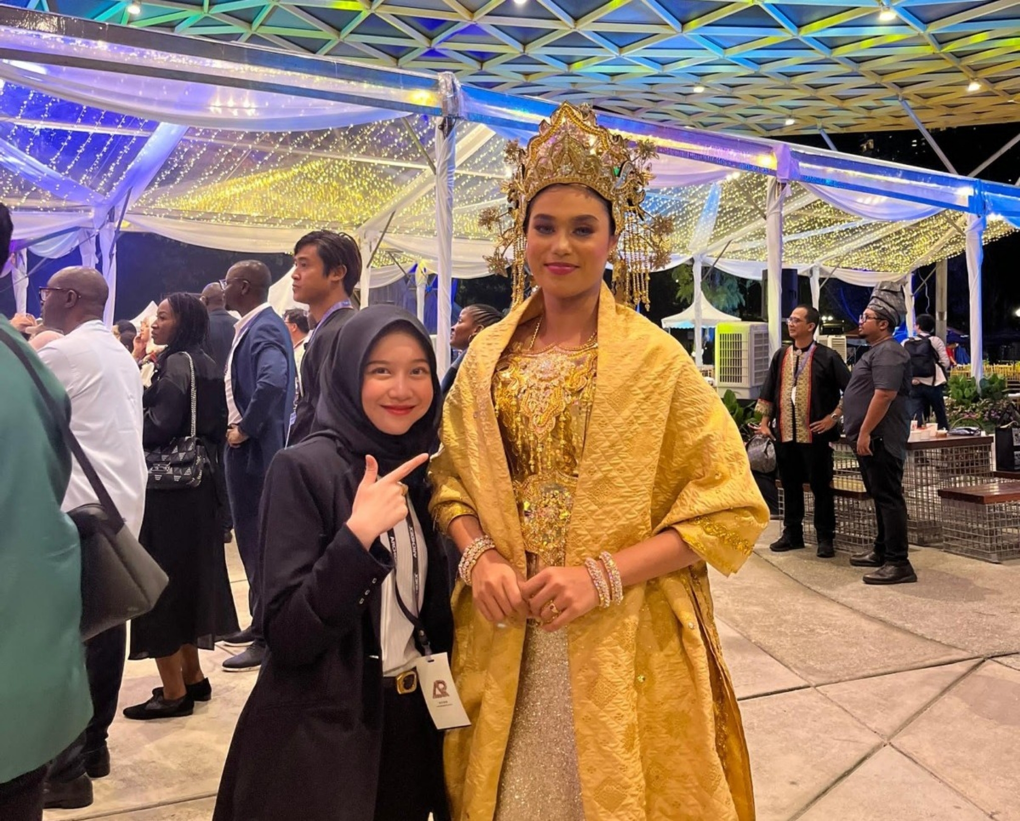 Young woman in black hijab smiling and pointing beside a tall person wearing ornate gold traditional dress and headdress.