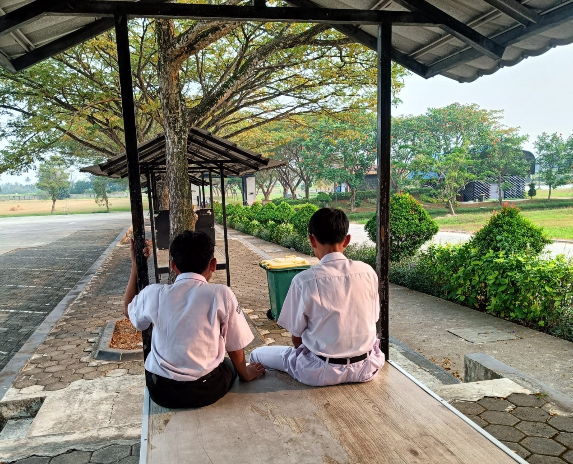 Two boys in white school uniforms sitting on a covered wooden platform, facing a tree-lined courtyard and pathway.