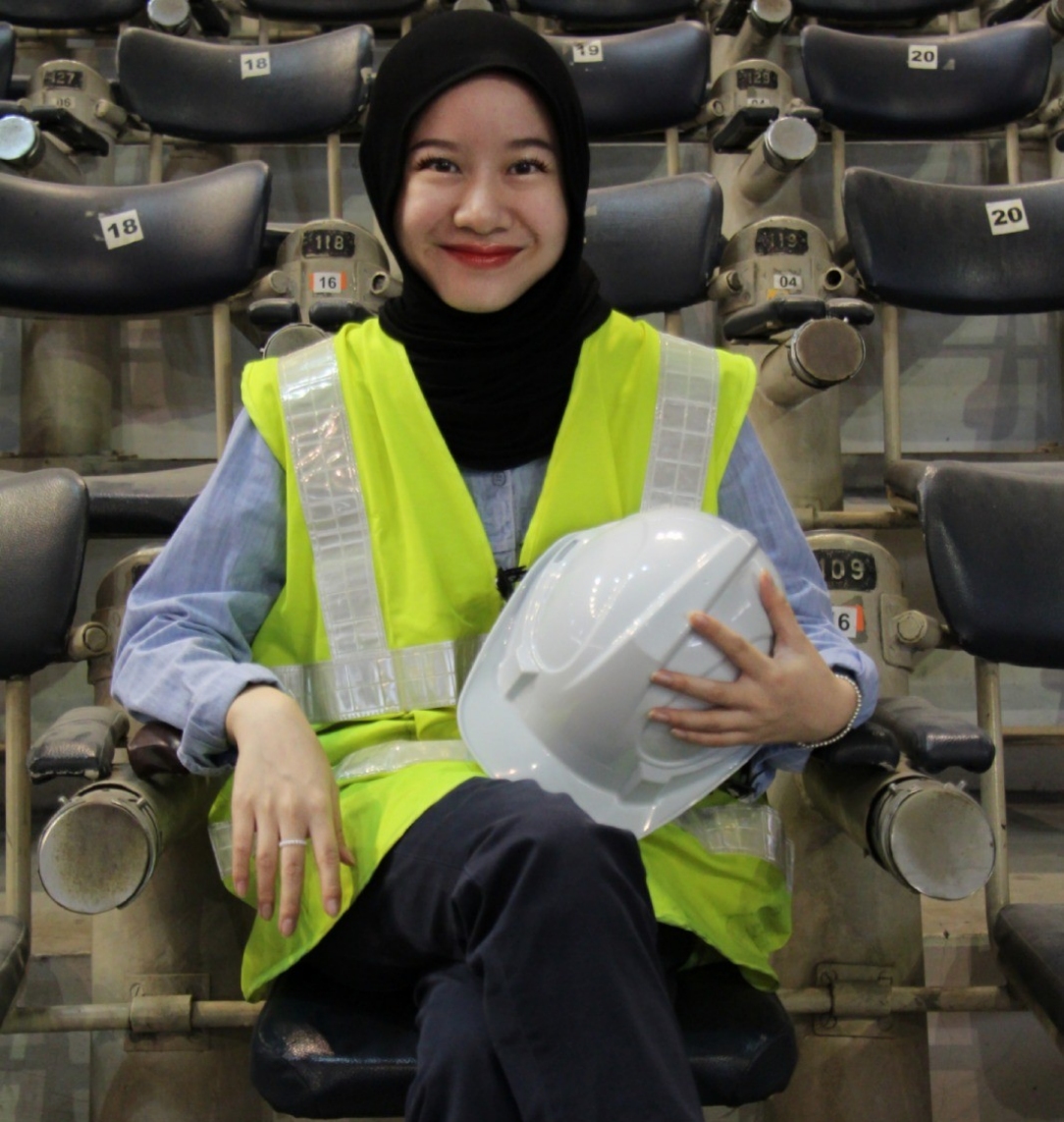 Smiling woman wearing a black hijab and yellow safety vest, seated in stadium-style seats and holding a white hard hat on her lap.
